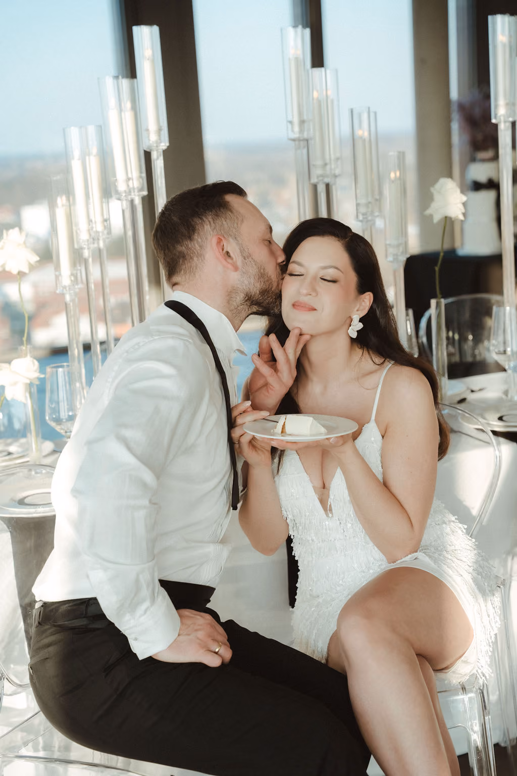 Ein Mann in weißem Hemd küsst eine lächelnde Frau in weißem Kleid auf die Wange, die ein Stück Kuchen auf einem Teller hält, vor einem festlich dekorierten Tisch mit Kerzen und weißen Rosen.