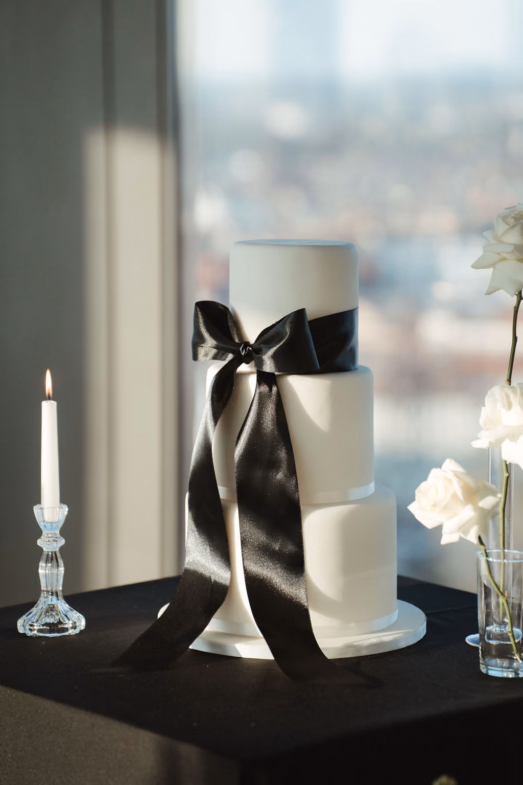 Weiße dreistöckige Hochzeitstorte mit einer großen schwarzen Schleife auf der mittleren Schicht, stehend auf einem schwarzen Tischtuch neben einer brennenden weißen Kerze und weißen Rosen in Glasvasen.