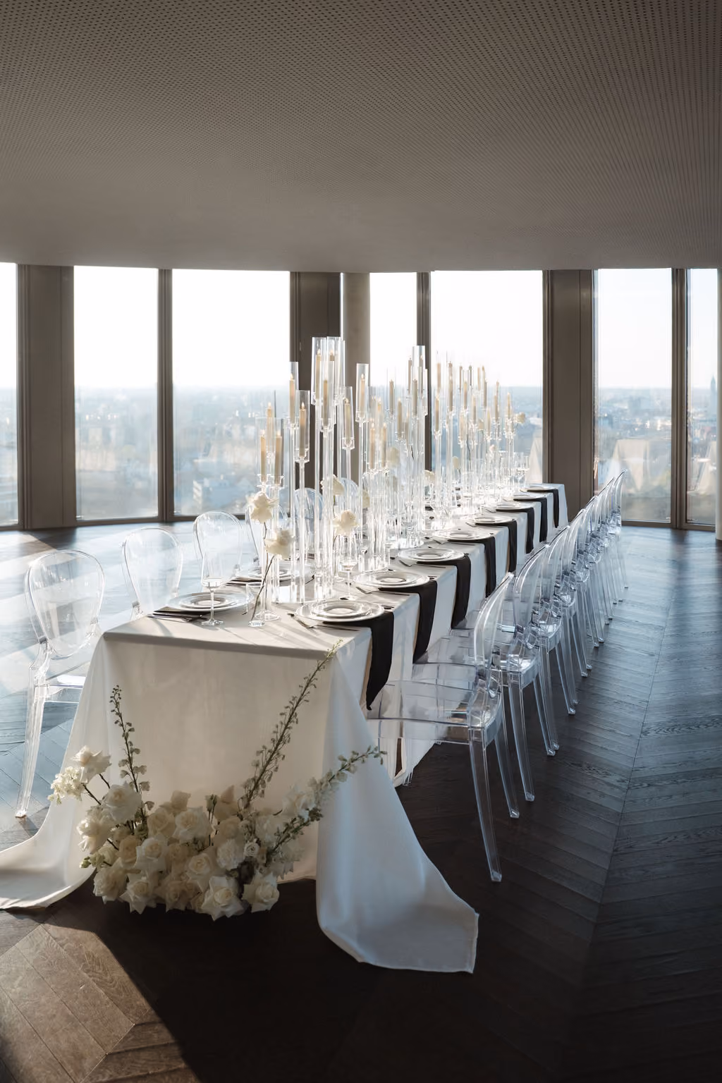 Eleganter, langer Esstisch in einem hochgelegenen Raum mit großen Fenstern und Stadtblick, gedeckt mit weißen Tischdecken, schwarzen Läufern, transparenten Stühlen, hohen Kerzenhaltern und weißen Rosen als Dekoration.