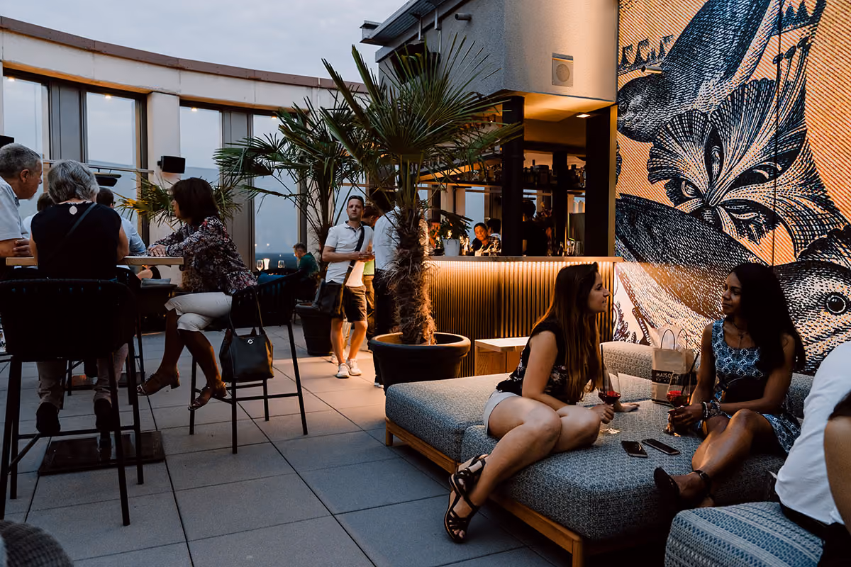 Zwei Frauen sitzen auf einem Sofa auf einer Dachterrasse mit Getränken in der Hand, vor einer Wand mit großem Vogelkunstwerk, während andere Gäste an Tischen und an der Bar im Hintergrund unter Pflanzen und Abendlicht entspannen.