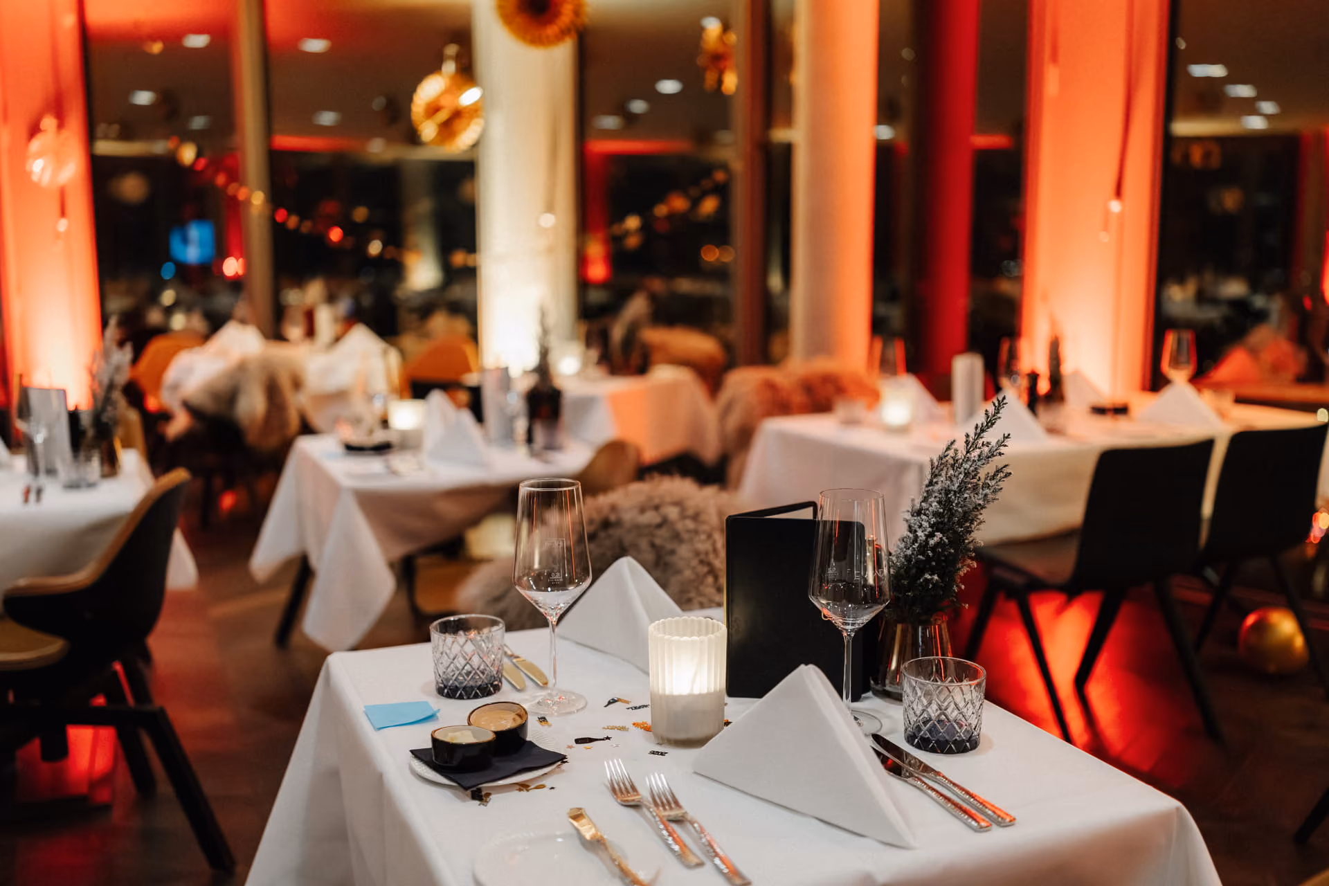 Elegantes Restaurant bei Kerzenlicht mit dekorierten Tischen, weißen Tischdecken, Weingläsern, Menükarten und sanfter roter Beleuchtung.