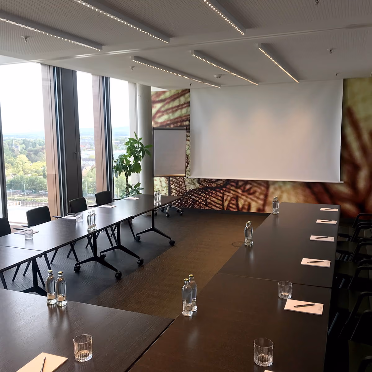 Moderner Konferenzraum mit großen Fenstern, zwei langen, U-förmig angeordneten dunklen Tischen, Stühlen, Wasserflaschen, Gläsern, Notizblöcken mit Stiften sowie einem Flipchart und einer großen weißen Leinwand an der Wand.
