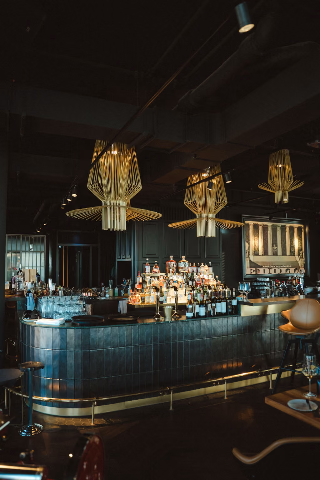 Moderne Bar mit dunkelblauen Fliesen, goldenen Design-Hängelampen über der Theke, zahlreichen Spirituosenflaschen, Barhockern und einem großen historischen Bild an der Wand.