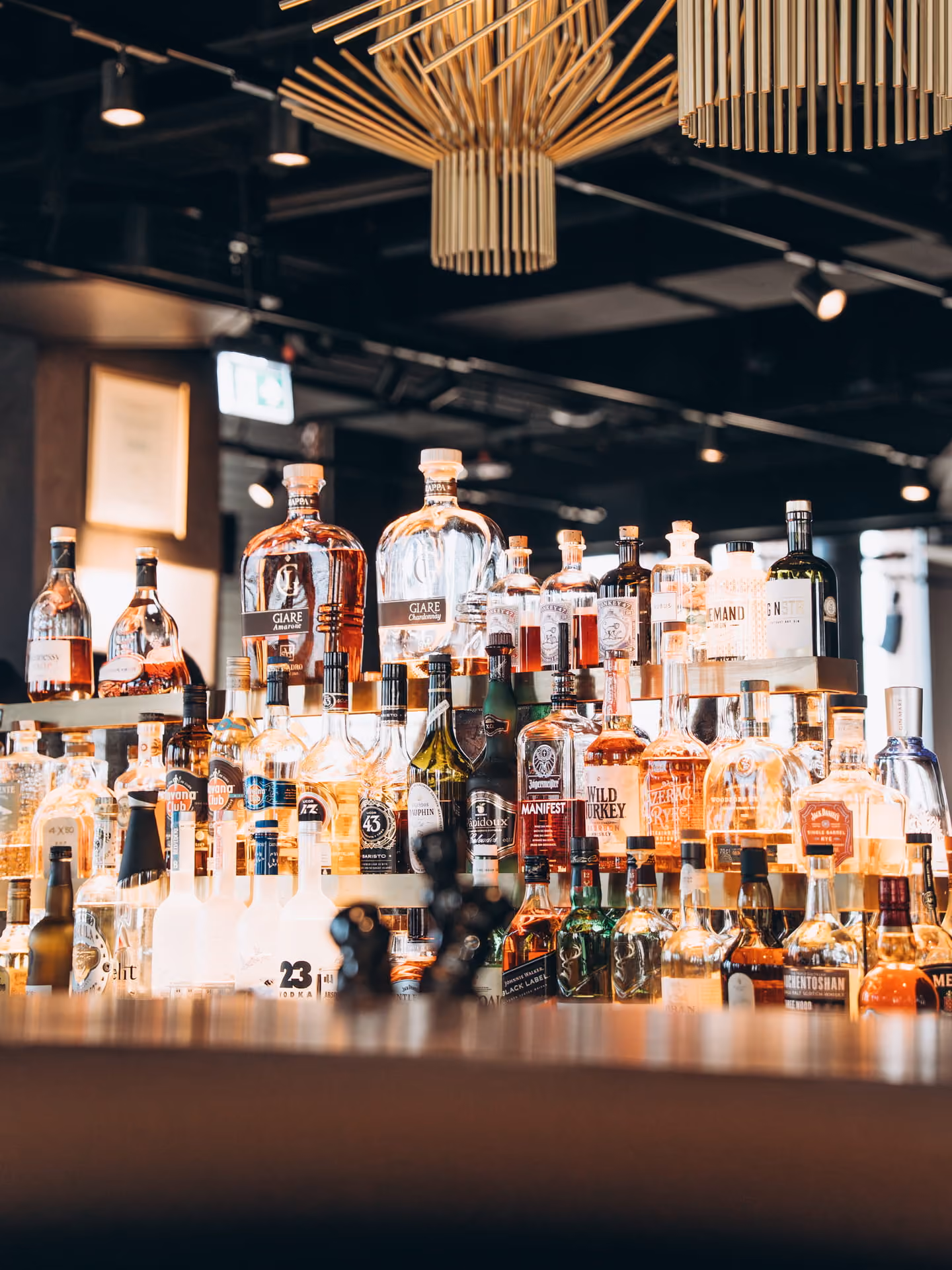 Regal mit verschiedenen Flaschen alkoholischer Getränke in einer Bar mit warmem Licht und modernen Holzlamellen-Deckenleuchten.