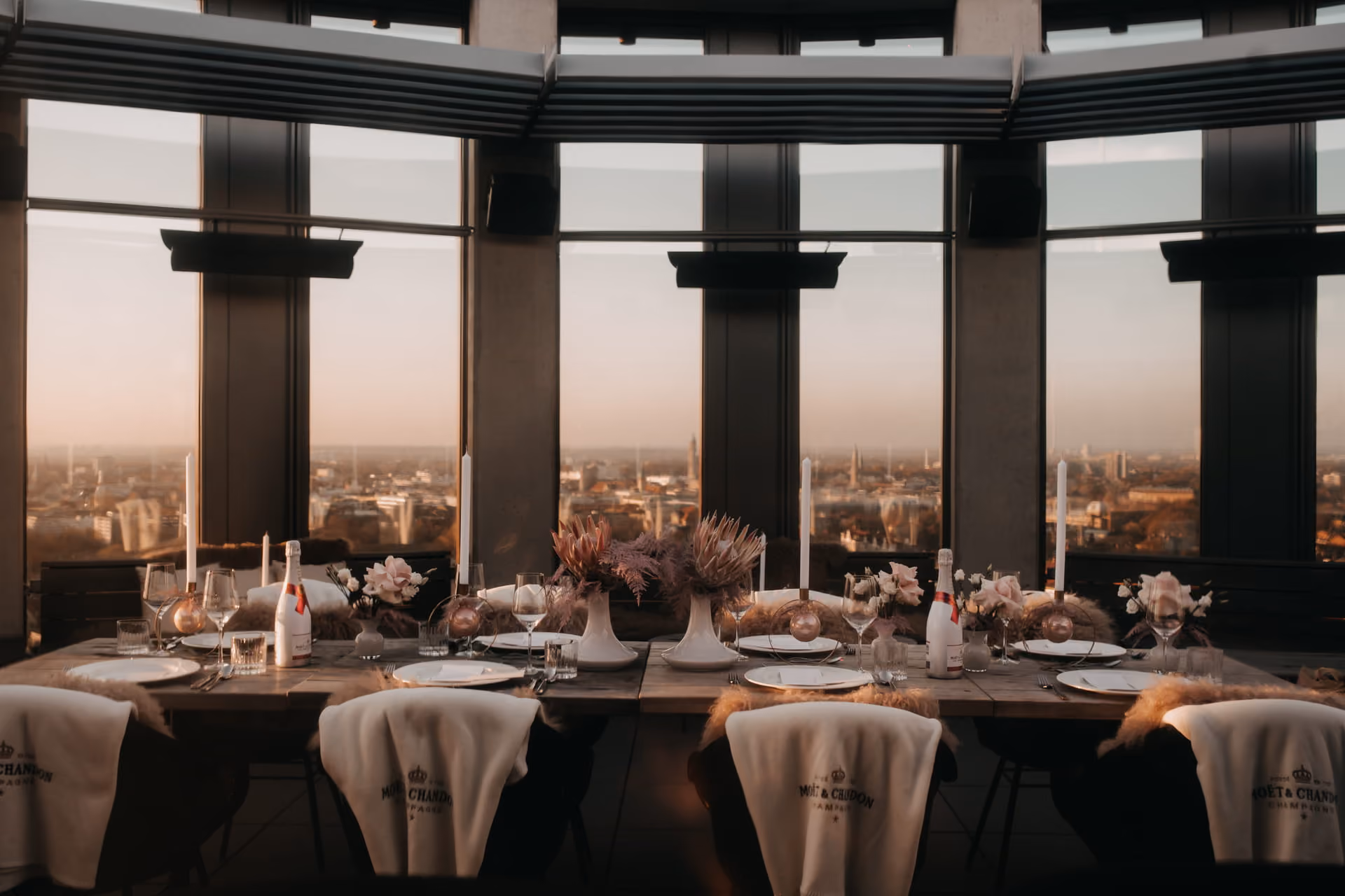 Eleganter gedeckter Tisch mit Kerzen, Champagnerflaschen, Blumenarrangements und weißen Stühlen mit Moët & Chandon Decken vor großen Fenstern mit Blick auf eine Stadt bei Sonnenuntergang.