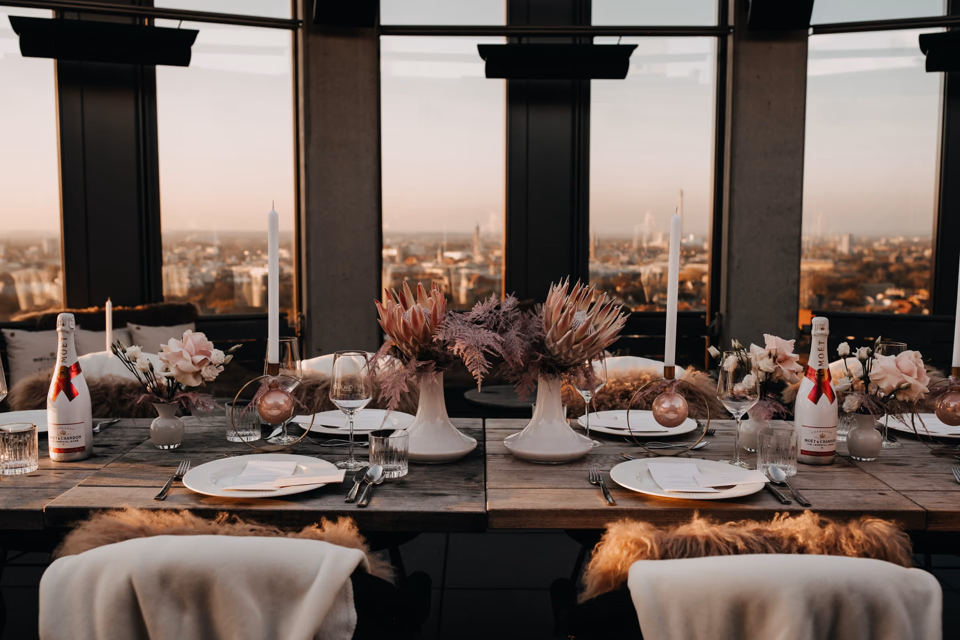 Eleganter eingedeckter Holztisch mit weißen Tellern, Weingläsern, modernen Blumenarrangements und Moët & Chandon Champagnerflaschen vor einem großen Fenster mit Stadtblick bei Sonnenuntergang.