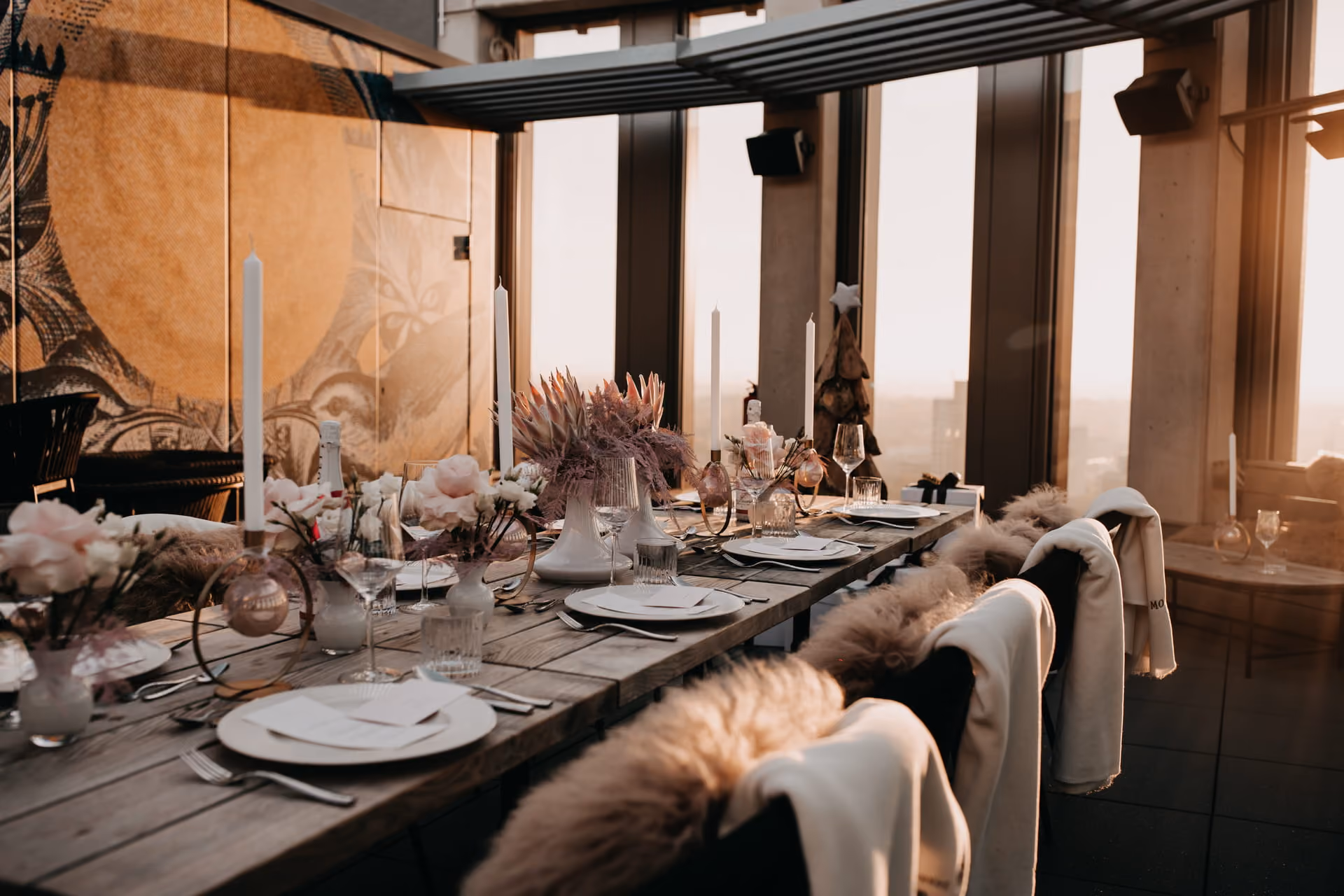 Eleganter gedeckter Holztisch mit weißen Kerzen, Blumenarrangements und Gläsern in einem modern eingerichteten Raum mit großen Fenstern und Blick auf die Stadt bei Sonnenuntergang.