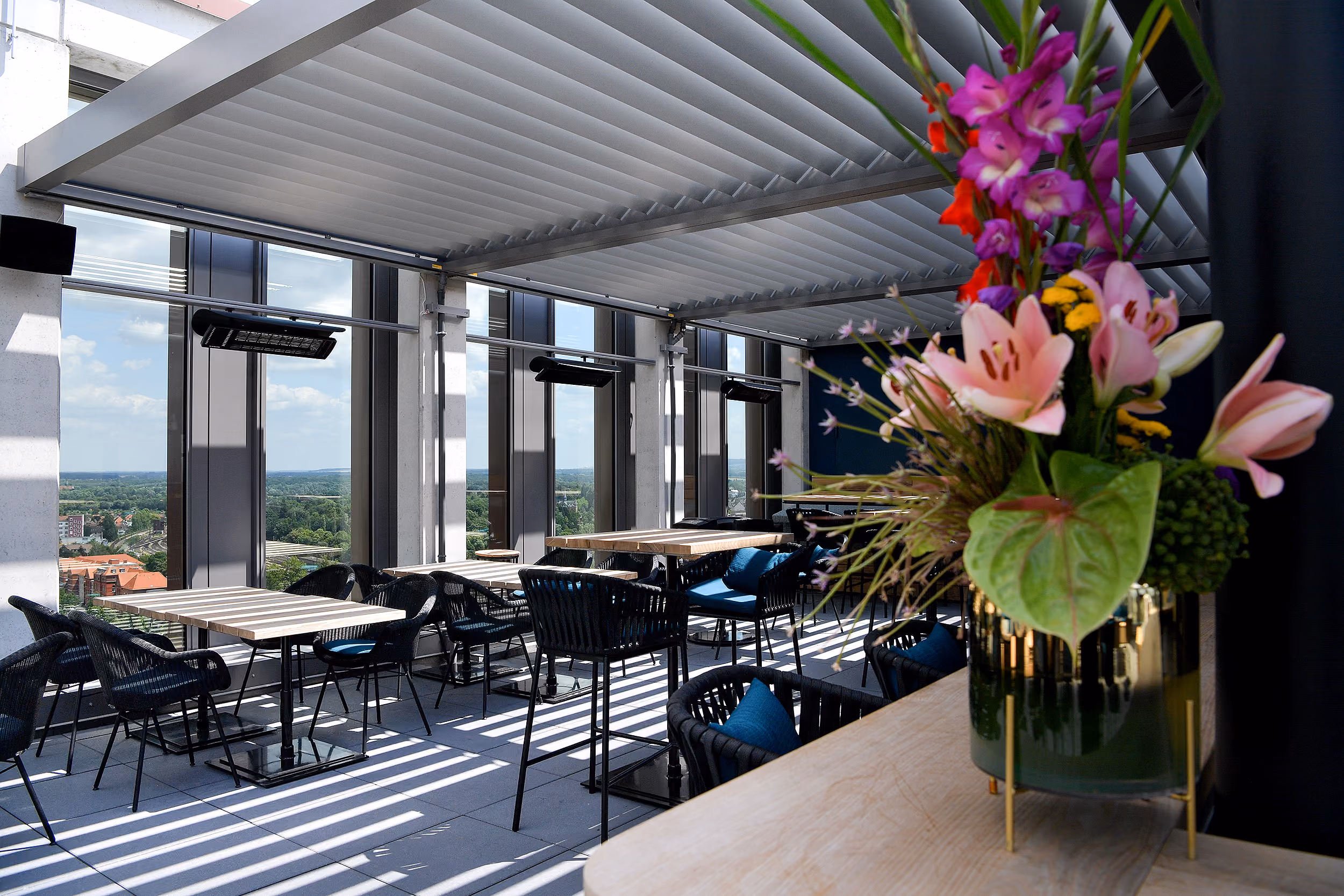 Moderne Dachterrasse mit Tischen und schwarzen Stühlen mit blauen Kissen, überdacht von einer lichten Lamellenstruktur, im Vordergrund ein Blumenstrauß mit pinken Lilien und anderen bunten Blumen in einem goldenen Vase.