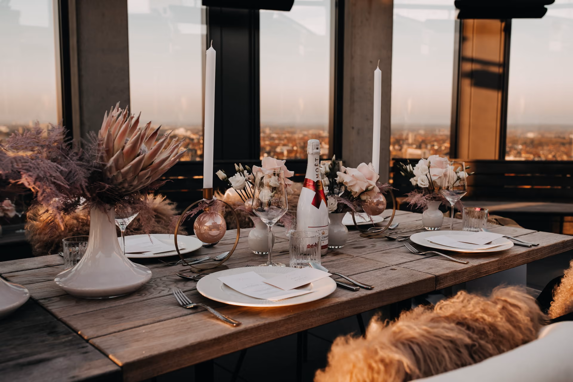 Eleganter Esstisch mit rustikalem Holz, gedeckt für zwei mit weißen Tellern, Besteck, Weingläsern und Menükarten, dekoriert mit weißen Kerzen, rosa Blumenarrangements und einer Flasche Moët Champagner, vor großen Fenstern mit Blick auf eine Stadtlandschaft bei Sonnenuntergang.