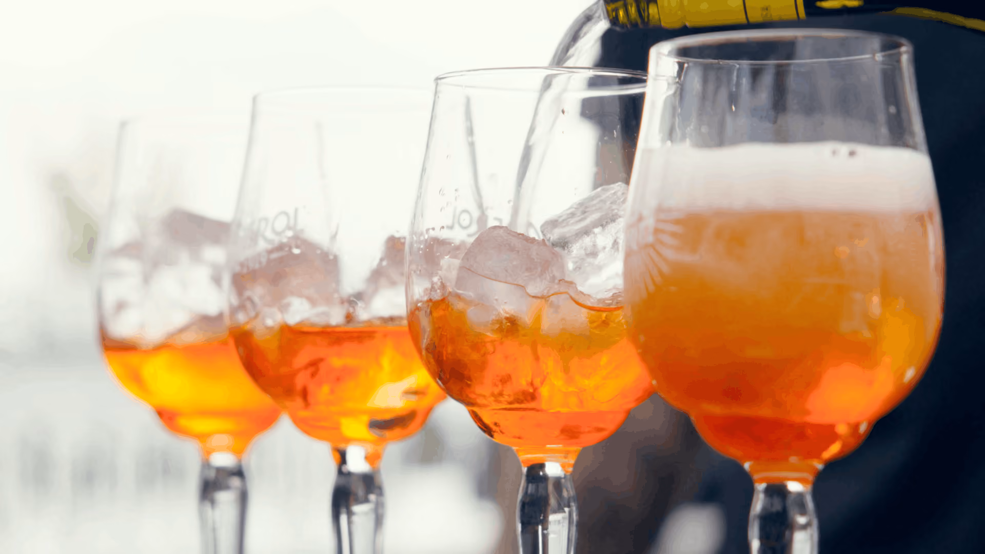 Vier Weingläser mit Eiswürfeln, die nacheinander mit einem orangefarbenen Getränk gefüllt werden, wobei das vorderste Glas bereits einen Schaumrand hat.