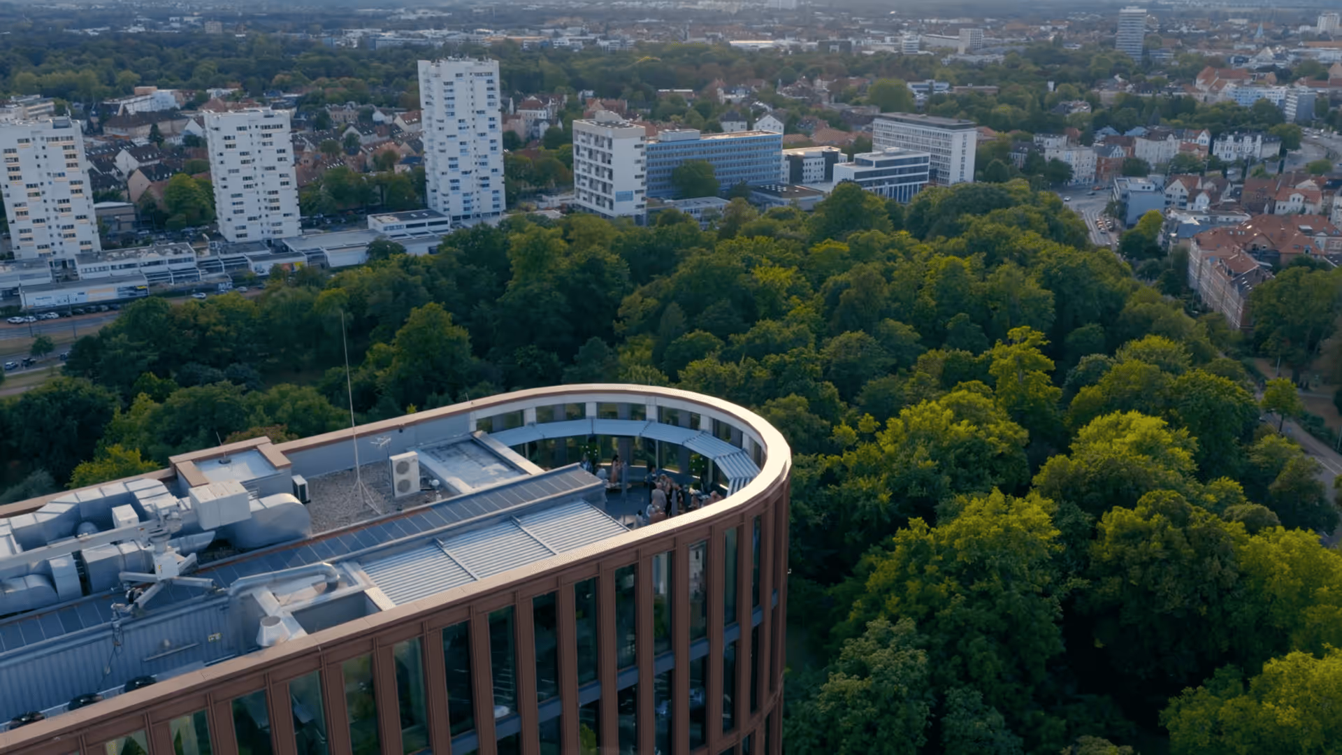 Luftaufnahme eines modernen, abgerundeten Bürogebäudes mit begrünter Umgebung aus Bäumen, im Hintergrund eine Stadt mit Wohn- und Bürogebäuden.