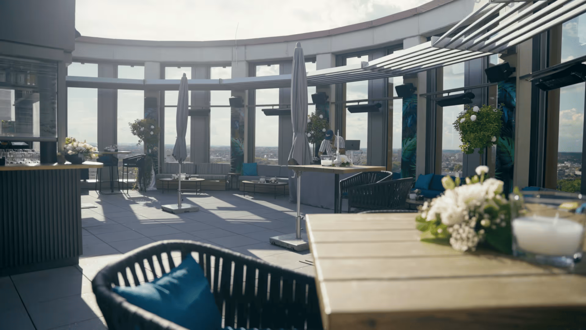 Dachterrasse mit modernem Mobiliar, einschließlich Tischen und Stühlen mit blauen Kissen, Sonnenschirmen und Pflanzen, mit einem Panoramablick auf die Stadt im Hintergrund.