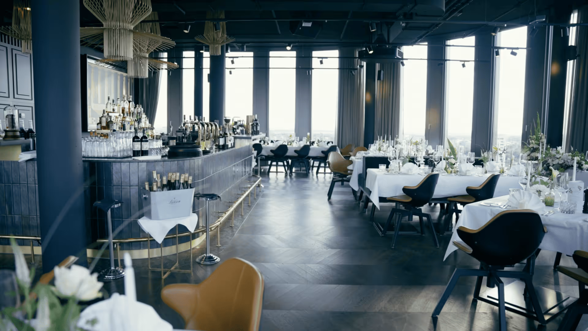 Modern, elegant restaurant interior with large windows letting in natural light, white tablecloths on tables set with glassware and floral centerpieces, and black chairs with tan cushions, alongside a dark tiled bar area with bottles and glassware.
