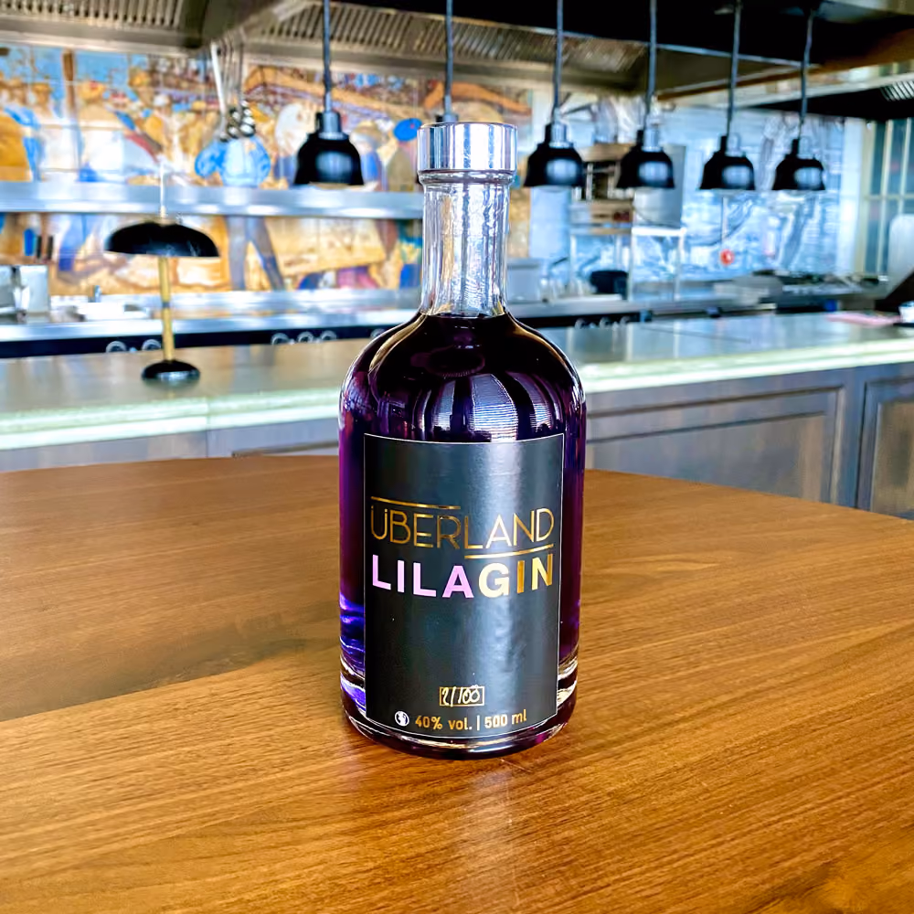 Flasche lila Gin mit schwarzem Etikett "Überland Lila Gin" und 40% Alkoholgehalt auf einem Holztisch in einer modernen Bar mit Lampen, Theke und Kunst im Hintergrund.