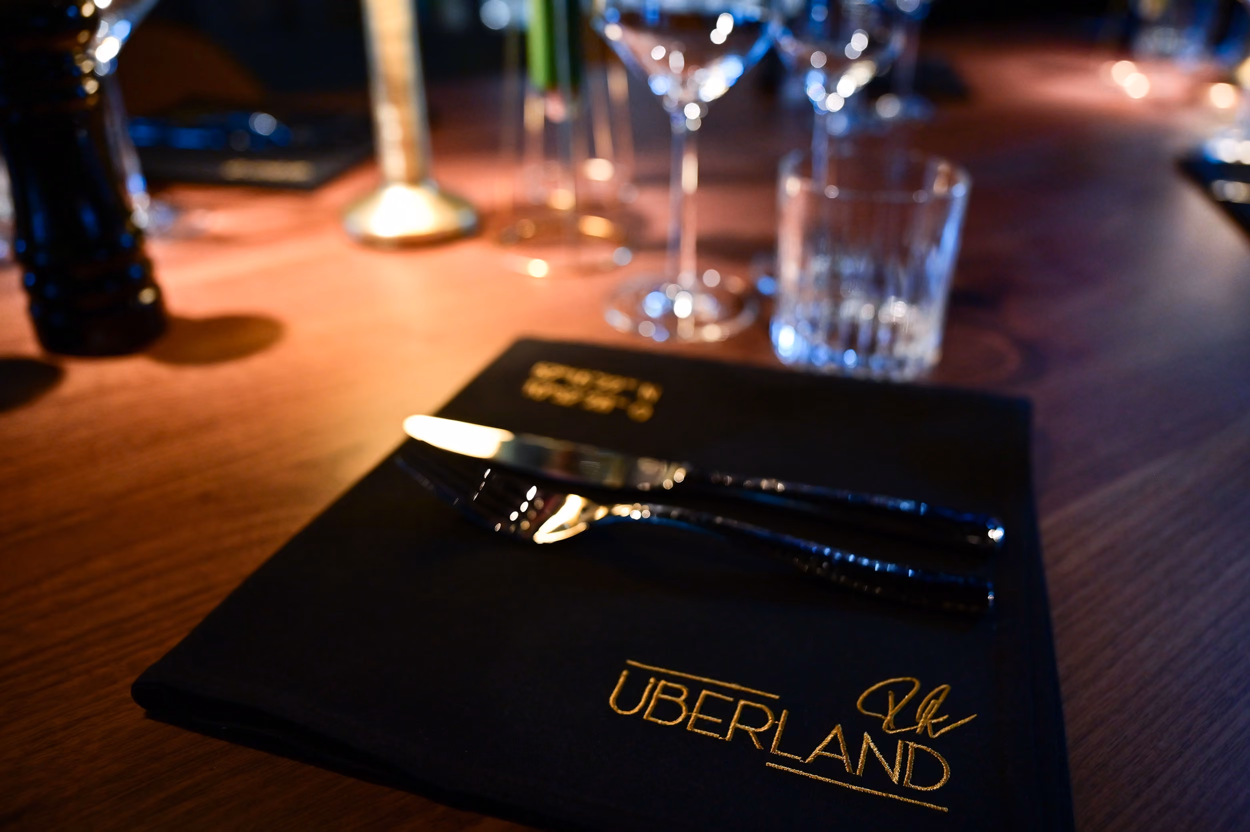 Auf einem Holztisch liegt eine schwarze Serviette mit der goldenen Aufschrift "ÜBERLAND", darauf liegen ein Messer und eine Gabel. Im Hintergrund stehen unscharf drei Weingläser, ein Wasserglas und ein Pfefferstreuer.