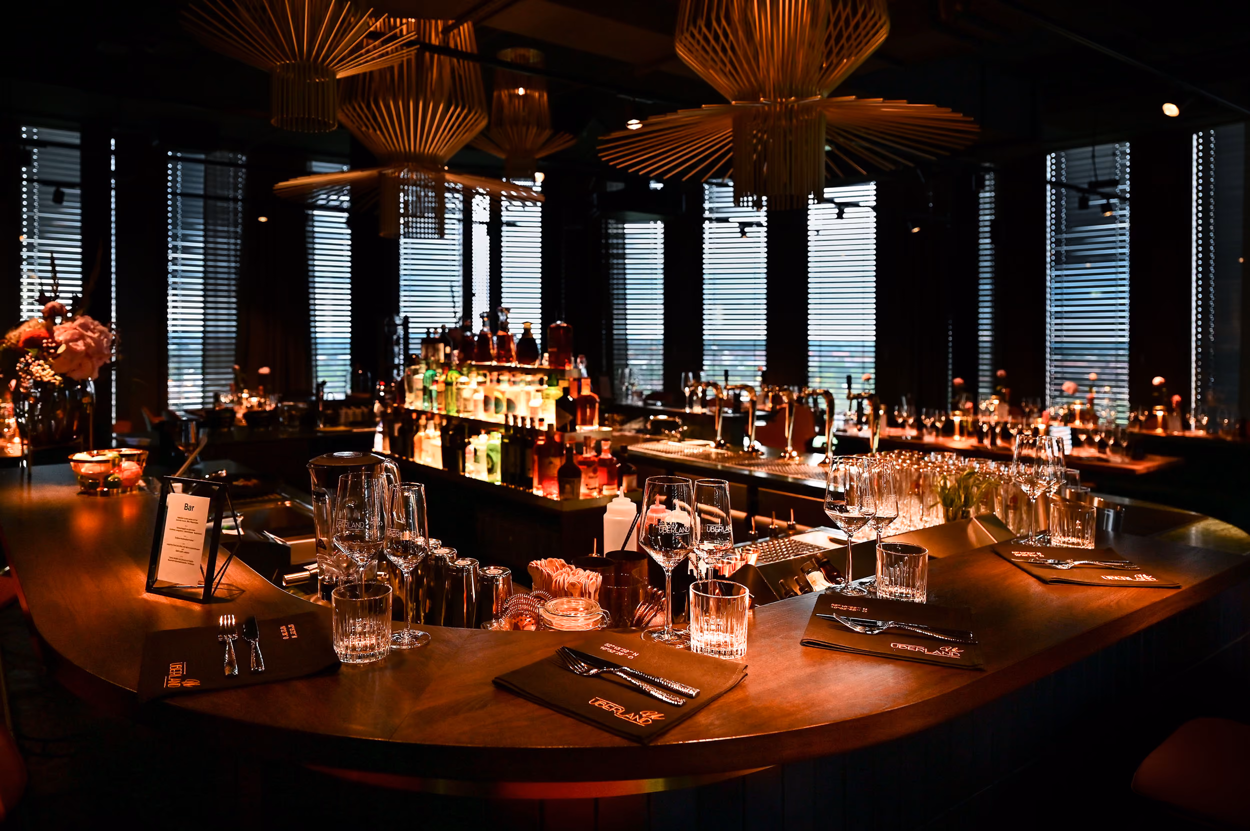 Elegante Bar mit U-förmigem Holzthekenbereich, dekoriert mit Weingläsern, Wassergläsern, Besteck auf Servietten und einer Auswahl an Flaschen beleuchtet im Hintergrund, mit großen Fenstern und modernen hängenden Lampen.