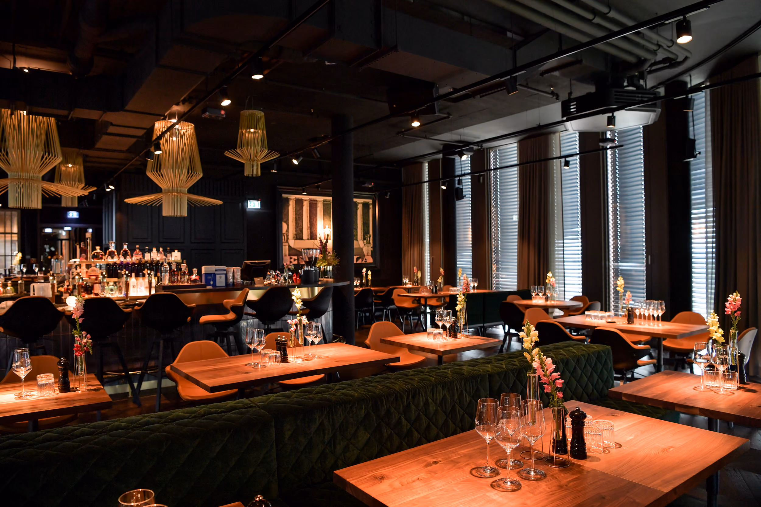 Elegantes Restaurantinnere mit modernen Holztischen, grünen samtigen Sitzbänken, orangen Stühlen und stilvollen Hängelampen über der Bar mit verschiedenen Flaschen, dekoriert mit kleinen Blumenvasen und Weingläsern auf den Tischen.