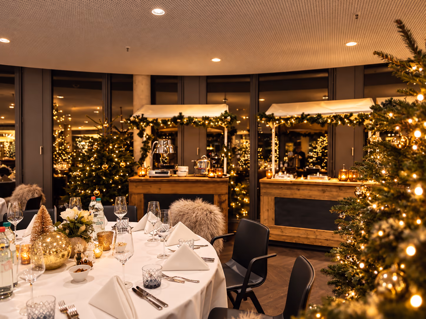 Festlich gedeckter runder Tisch mit weißen Servietten, Glas und Besteck in einem warm beleuchteten Raum mit Weihnachtsbäumen, dekoriert mit Lichterketten und goldenen Kugeln, im Hintergrund zwei Holzhütten mit Weihnachtsdekoration.