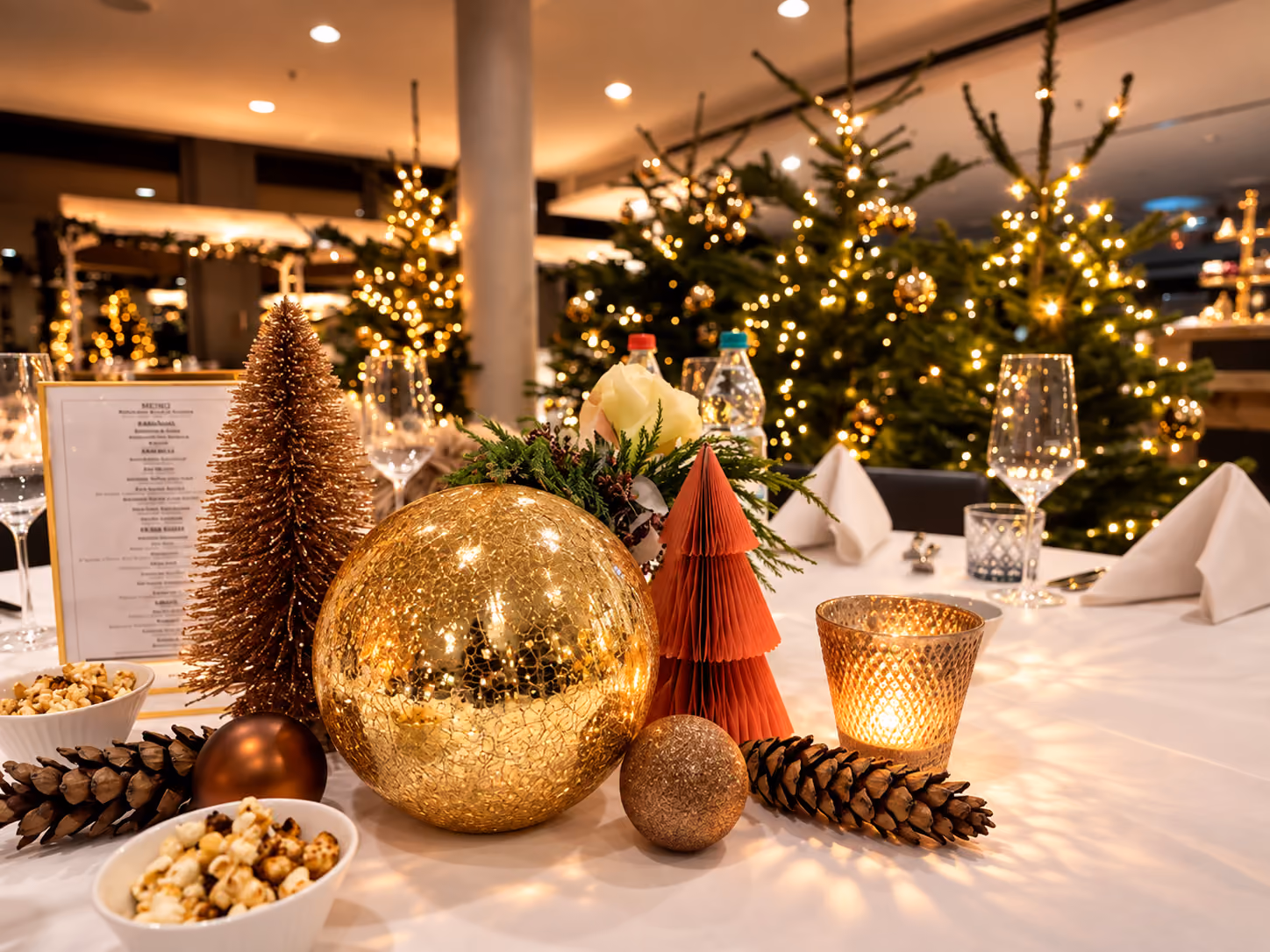 Weihnachtlich gedeckter Tisch mit goldener Glaskugel, kupferfarbenem Baum, roten Papierbäumchen, Zapfen und Kerzenlicht, im Hintergrund festlich beleuchtete Tannenbäume.