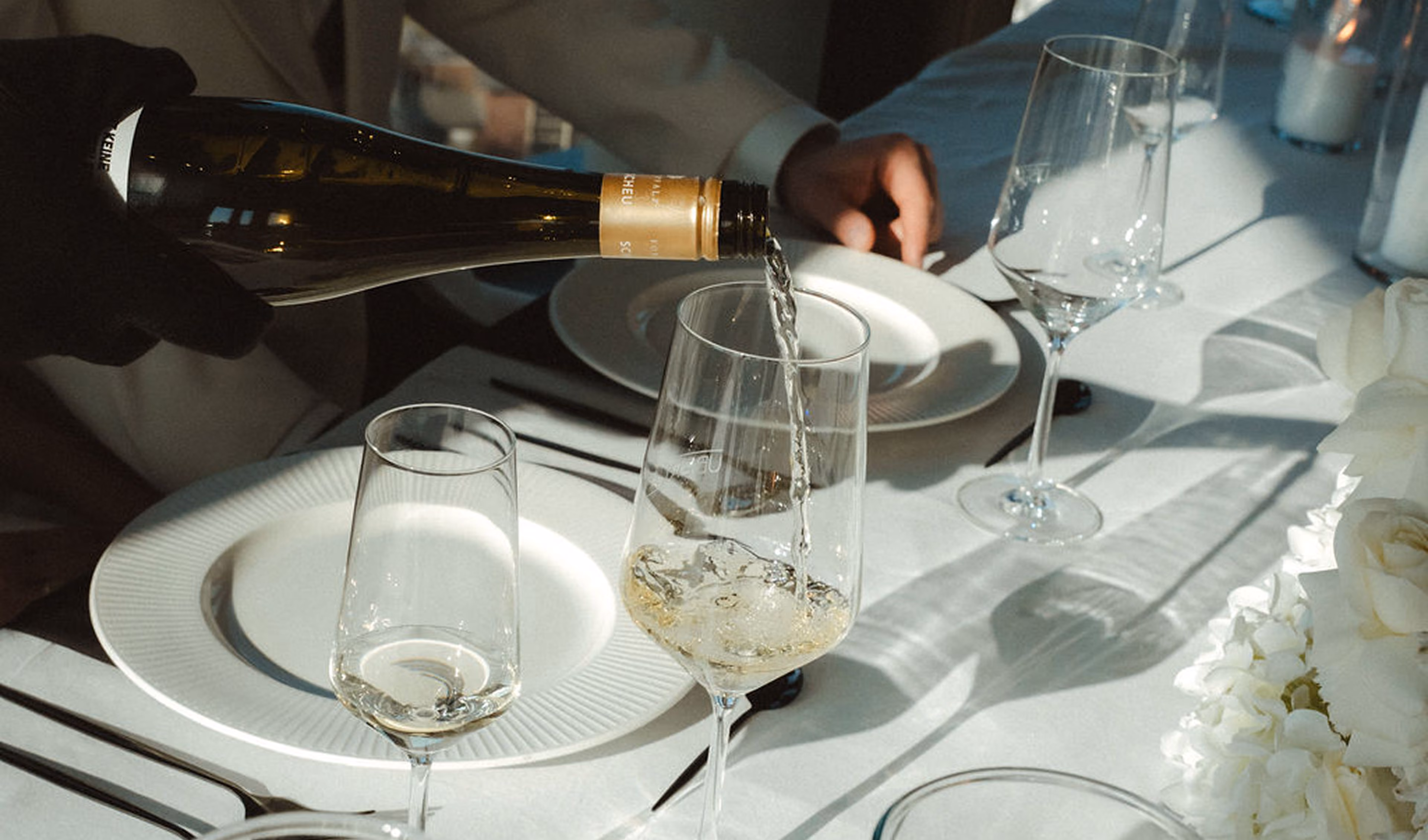 Person gießt weißen Wein aus einer Flasche in ein Weinglas auf einem gedeckten Tisch mit weißen Tellern, weiteren Gläsern und weißen Blumen.