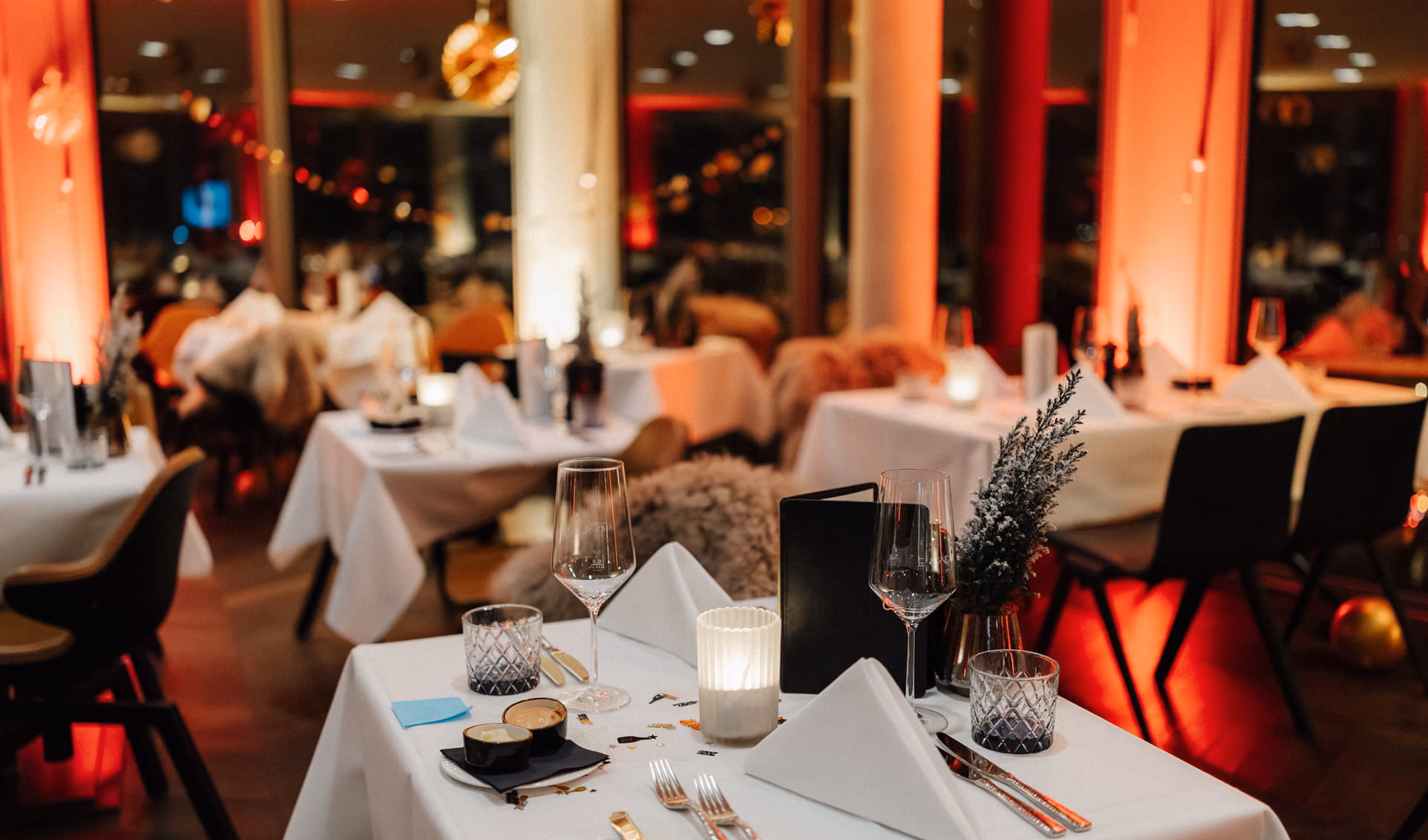 Weihnachts-Dinnerszene in einem festlich beleuchteten Restaurant mit weißen Tischdecken, Wein- und Wassergläsern, Kerzenlicht und dekorativem Grün auf den Tischen.