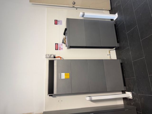 Two large gray battery storage units with warning signs on a beige wall in a room with black tiled floor, separated by white protective posts.