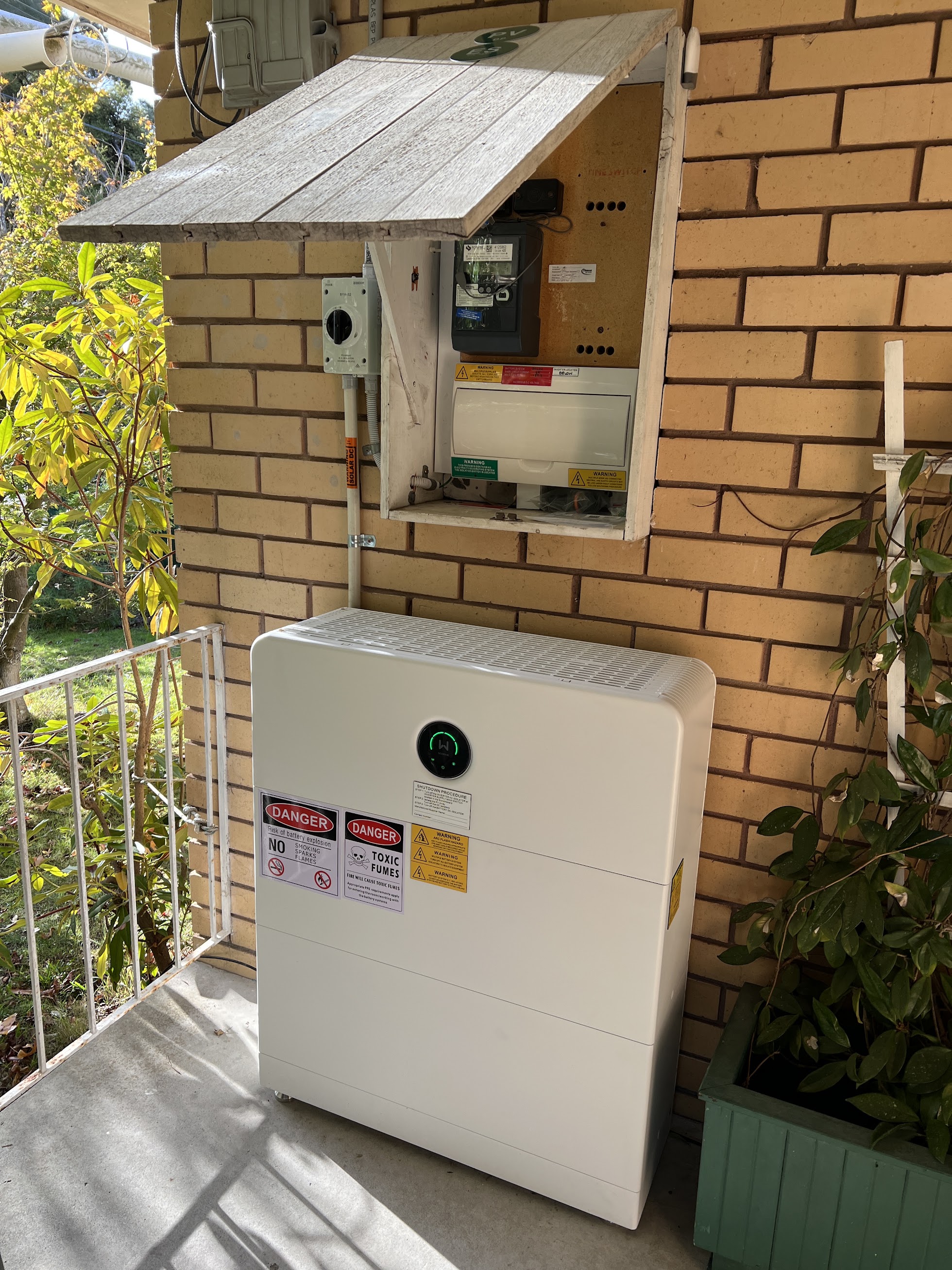 White electrical or utility box with multiple warning labels, including danger of battery explosion and toxic fumes, mounted outdoors against a brick wall under a small wooden awning, next to a green planter with foliage and a white metal railing.
