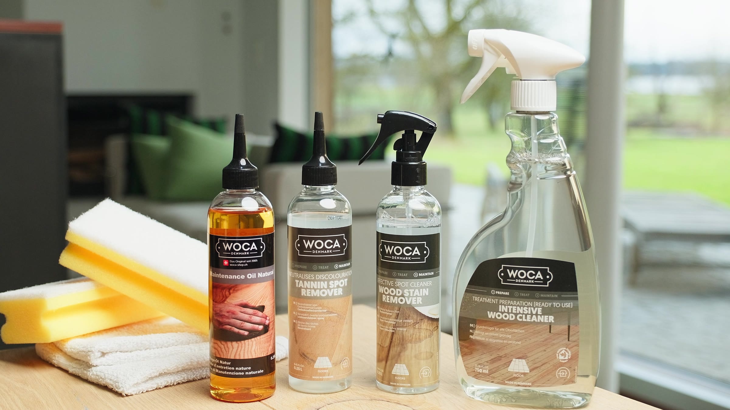 WOCA-Pflegeprodukte für Flecken auf einem Holztisch, dahinter ein helles Wohnzimmer