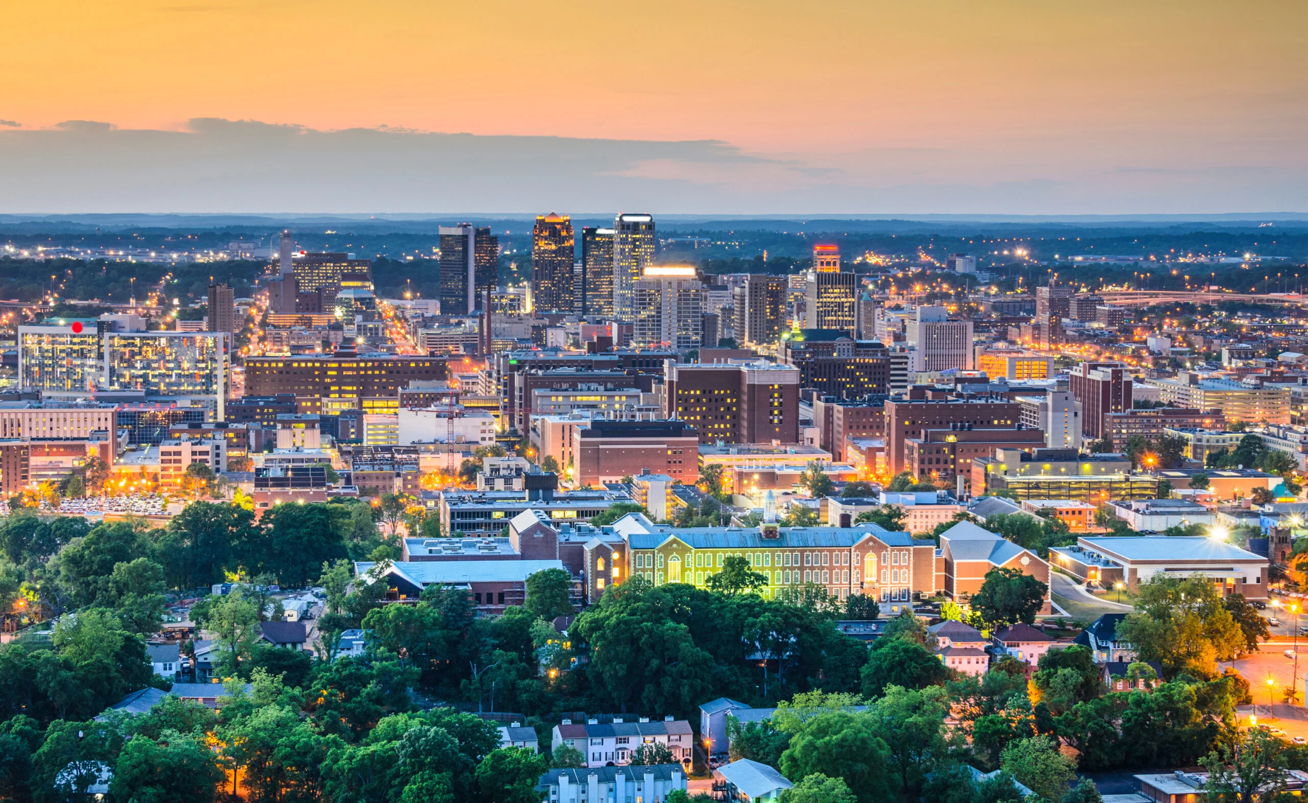 City skyline at dusk with illuminated buildings, streets, and a foreground of dense green trees and houses.