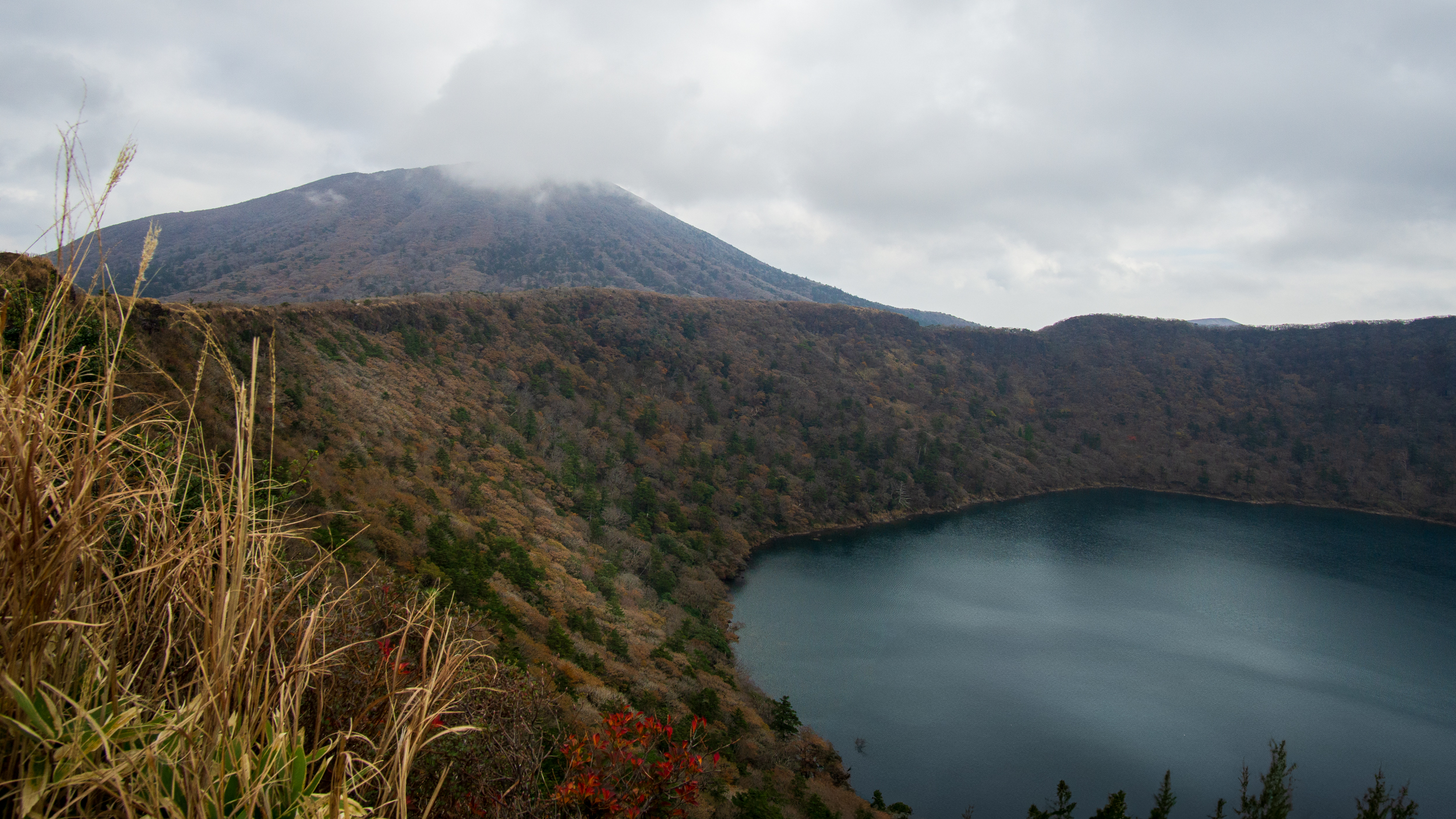 Kratersee unter einem wolkenverhangenen Vulkangipfel, im Vordergrund trockenes Gras