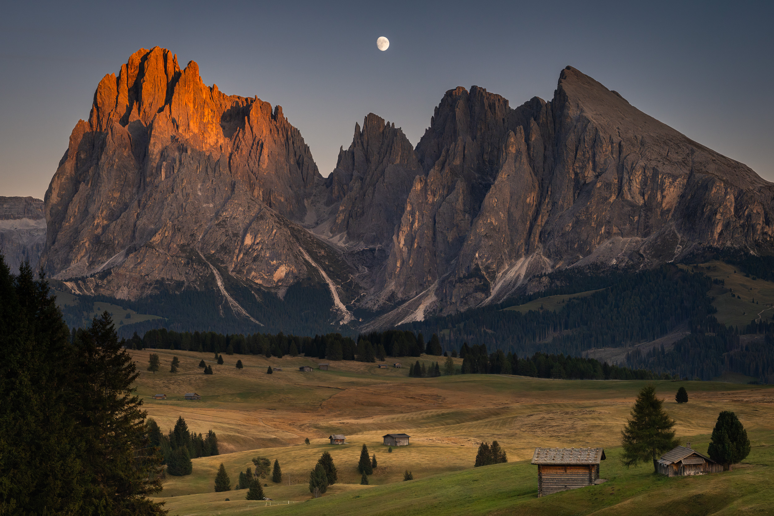 Panorama der Langkofelgruppe im Alpenglühen, darüber der Mond, davor Almwiesen und Hütten