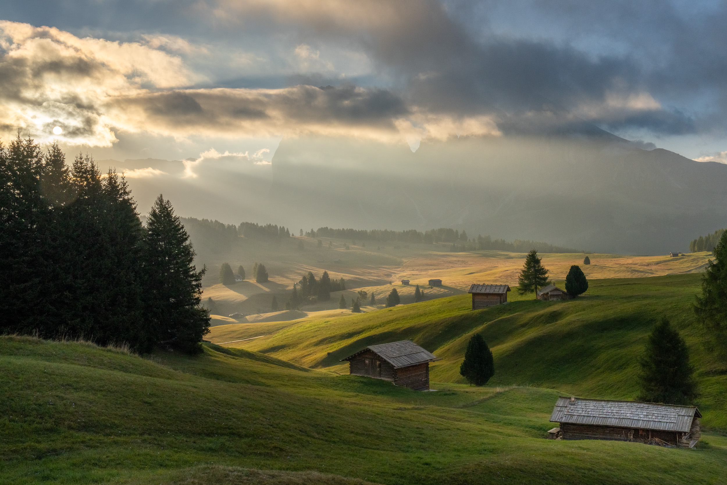Lichtstrahlen brechen durch Wolken über sanften Almwiesen mit Holzhütten