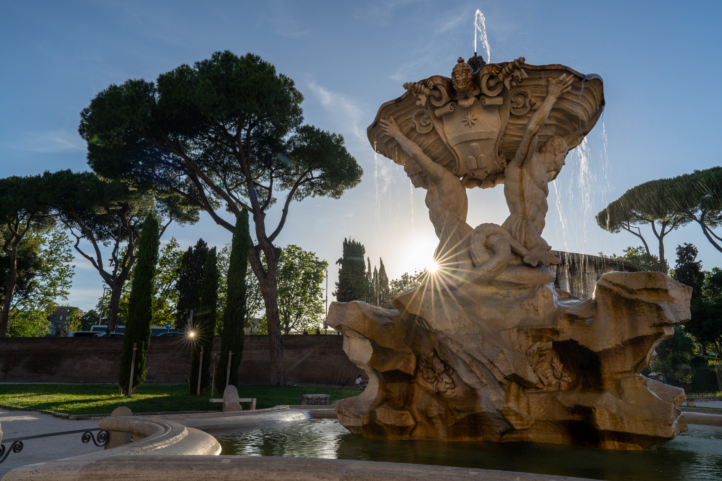 Tritonenbrunnen im Gegenlicht mit Sonnenstern, dahinter Pinien