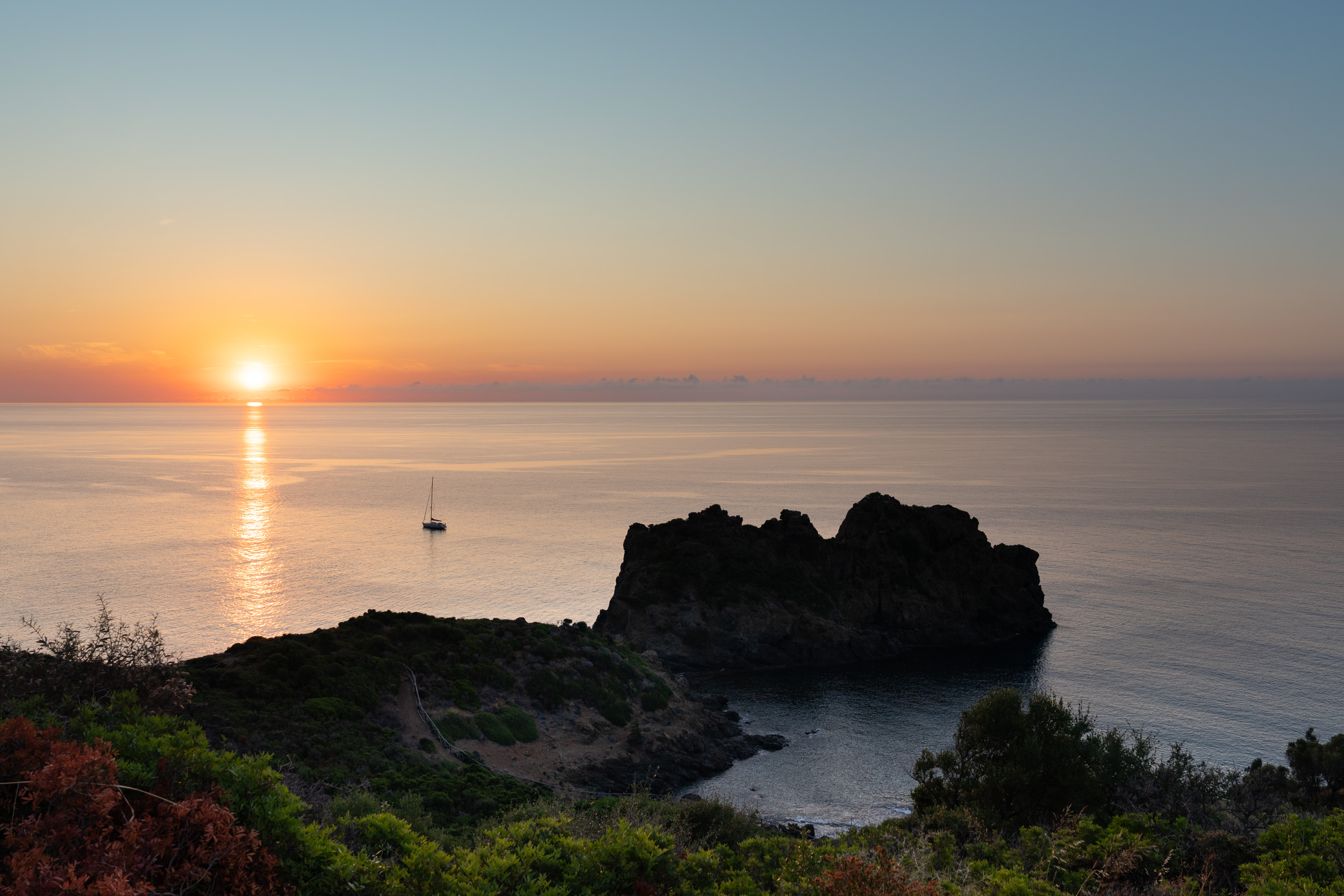 Sonnenaufgang über dem Meer, davor ein Felsen in einer kleinen Bucht und ein Segelboot