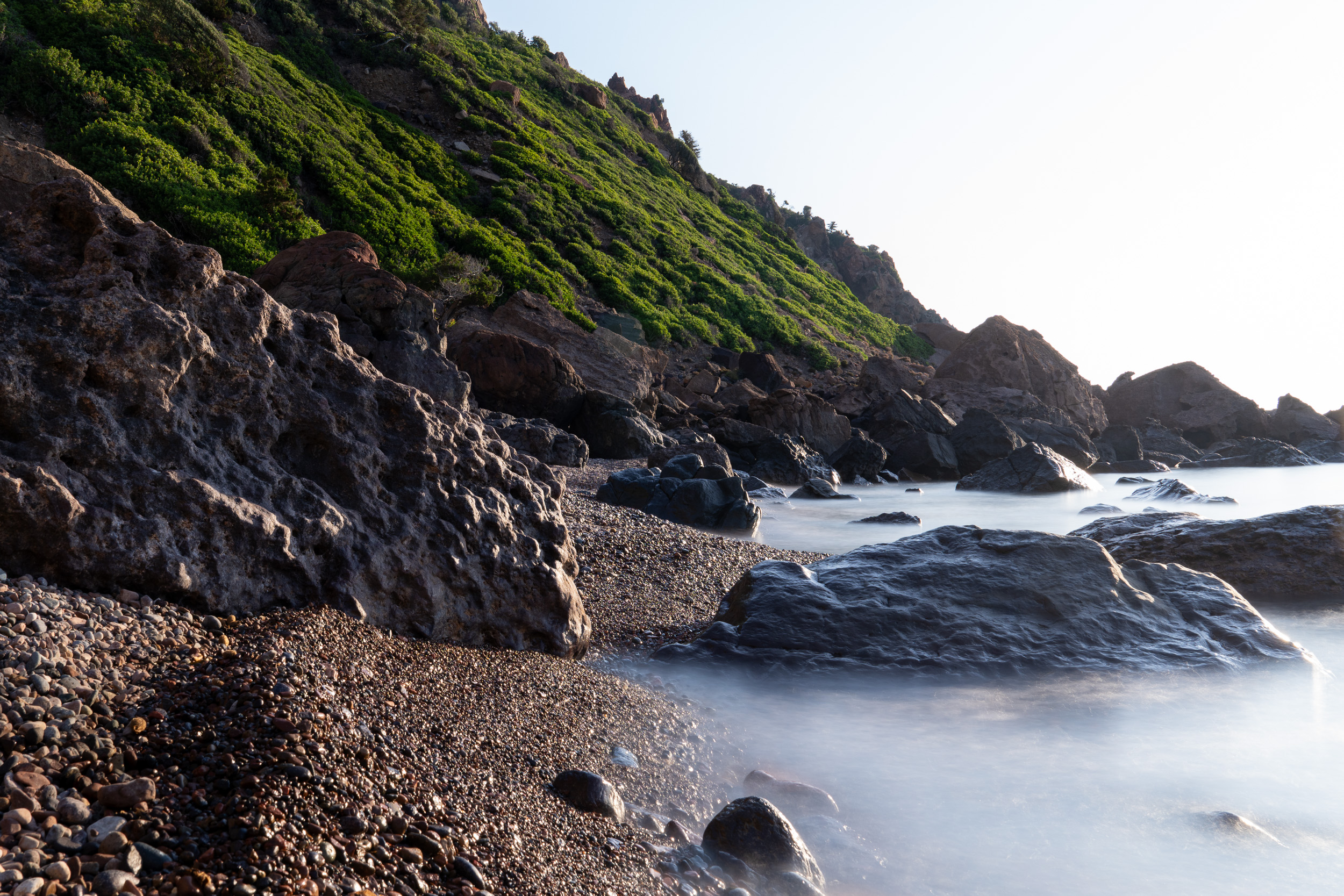Kieselstrand und dunkle Felsen im weichen Morgenlicht, das Wasser durch Langzeitbelichtung geglättet