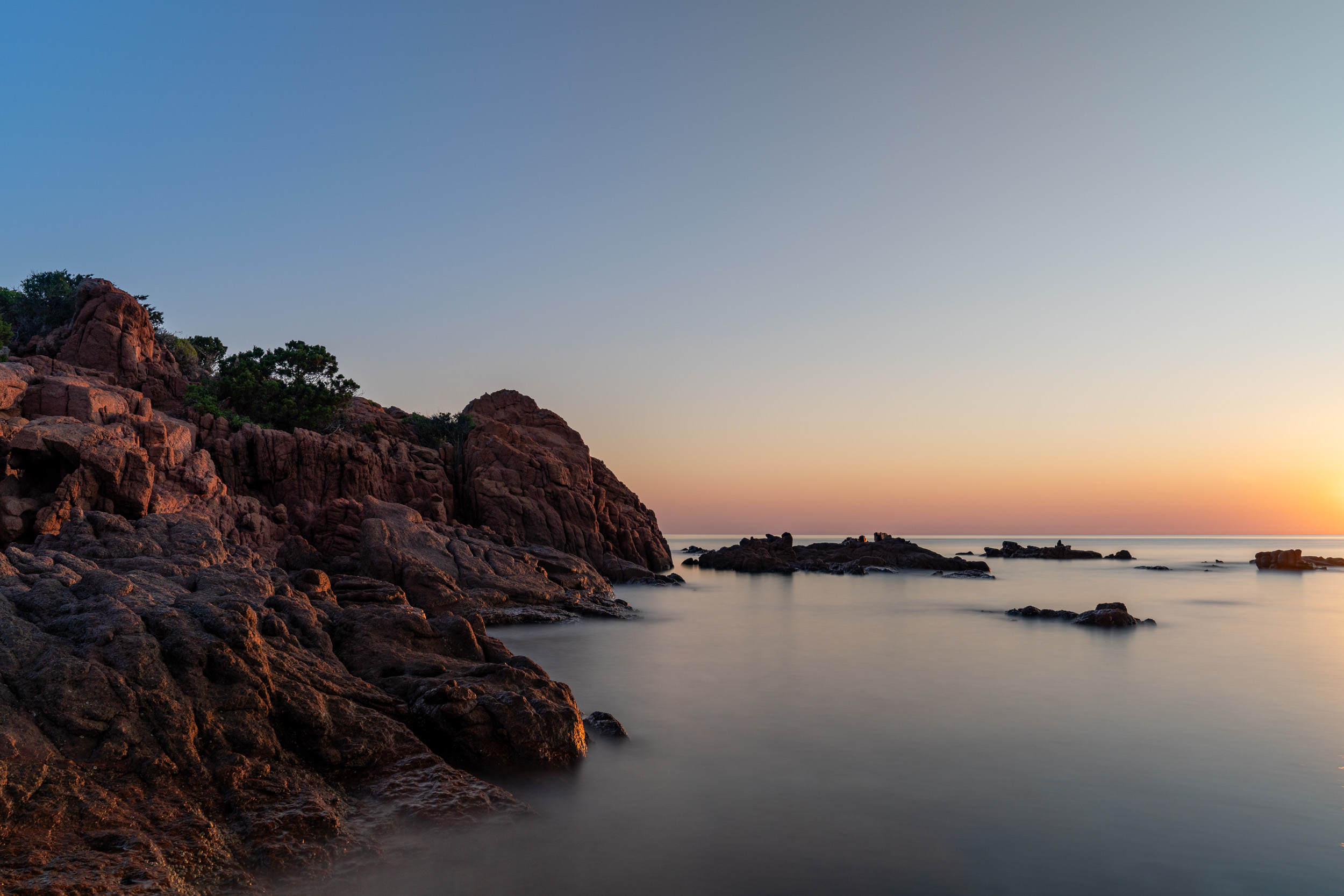 Rötliche Felsen an der Küste bei Sonnenaufgang über ruhigem Meer
