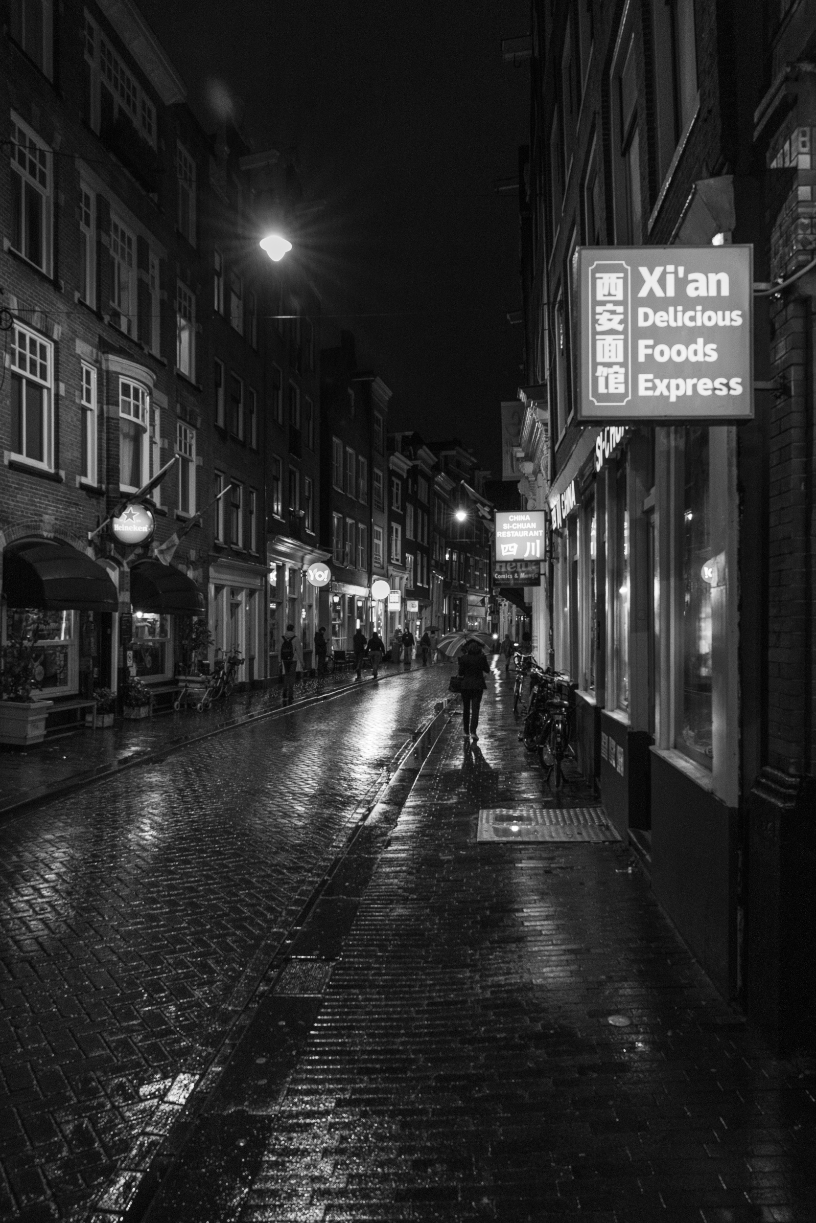 Nasse Gasse bei Nacht mit Passanten unter Regenschirmen, in Schwarzweiß