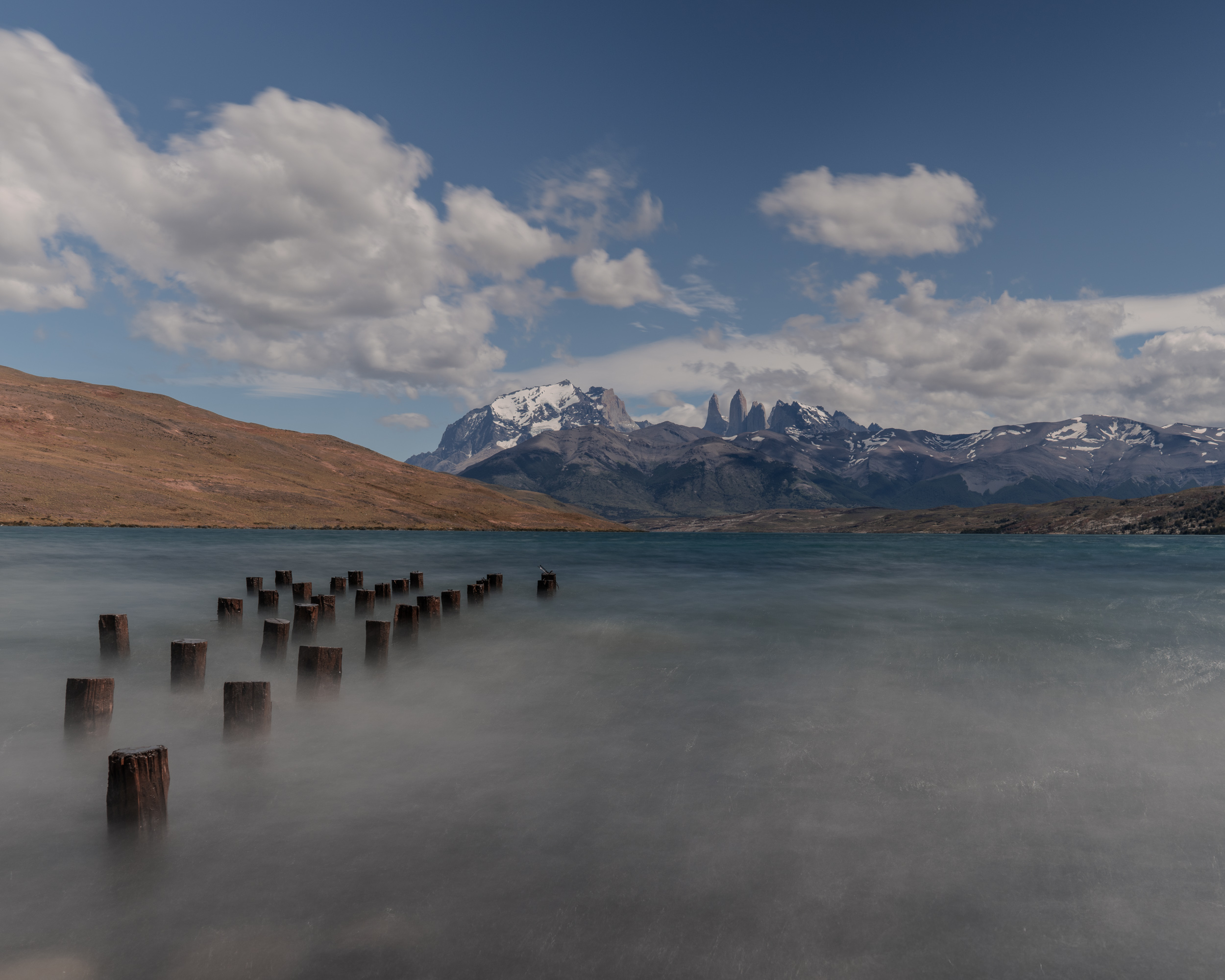 Alte Holzpfähle im See, dahinter schneebedeckte Berge
