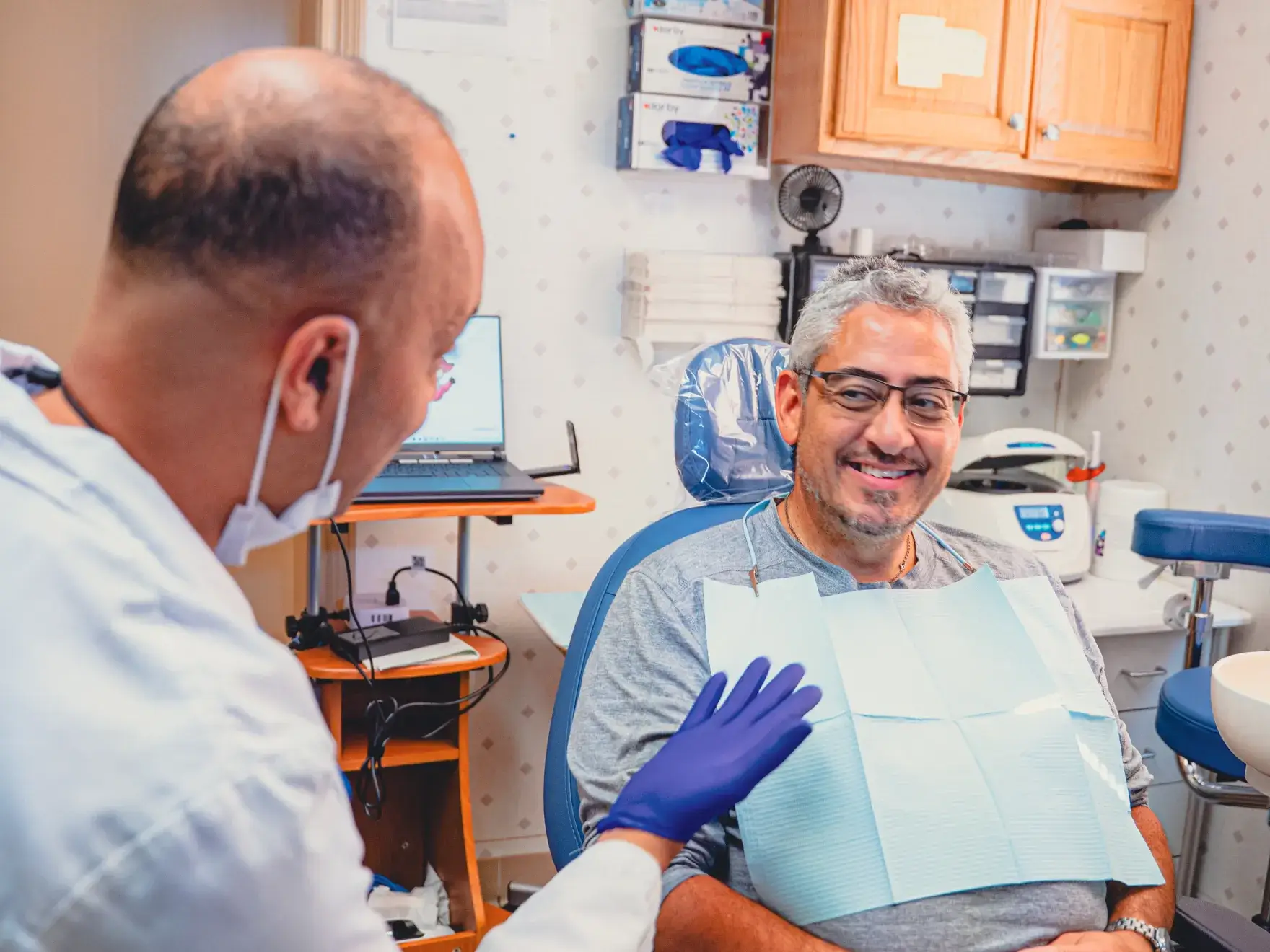 Dentist wearing gloves interacts with a smiling male patient seated in a dental chair in a clinic.