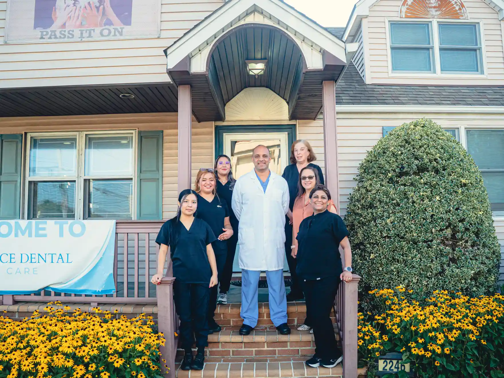 Dental care team posing outside a clinic entrance with a welcome banner and flower beds.