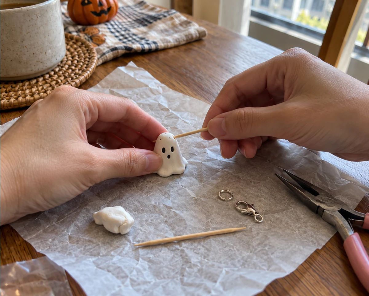 Hands pressing a face into a white polymer clay ghost with a toothpick on parchment paper, with a jump ring, lobster clasp and pliers nearby