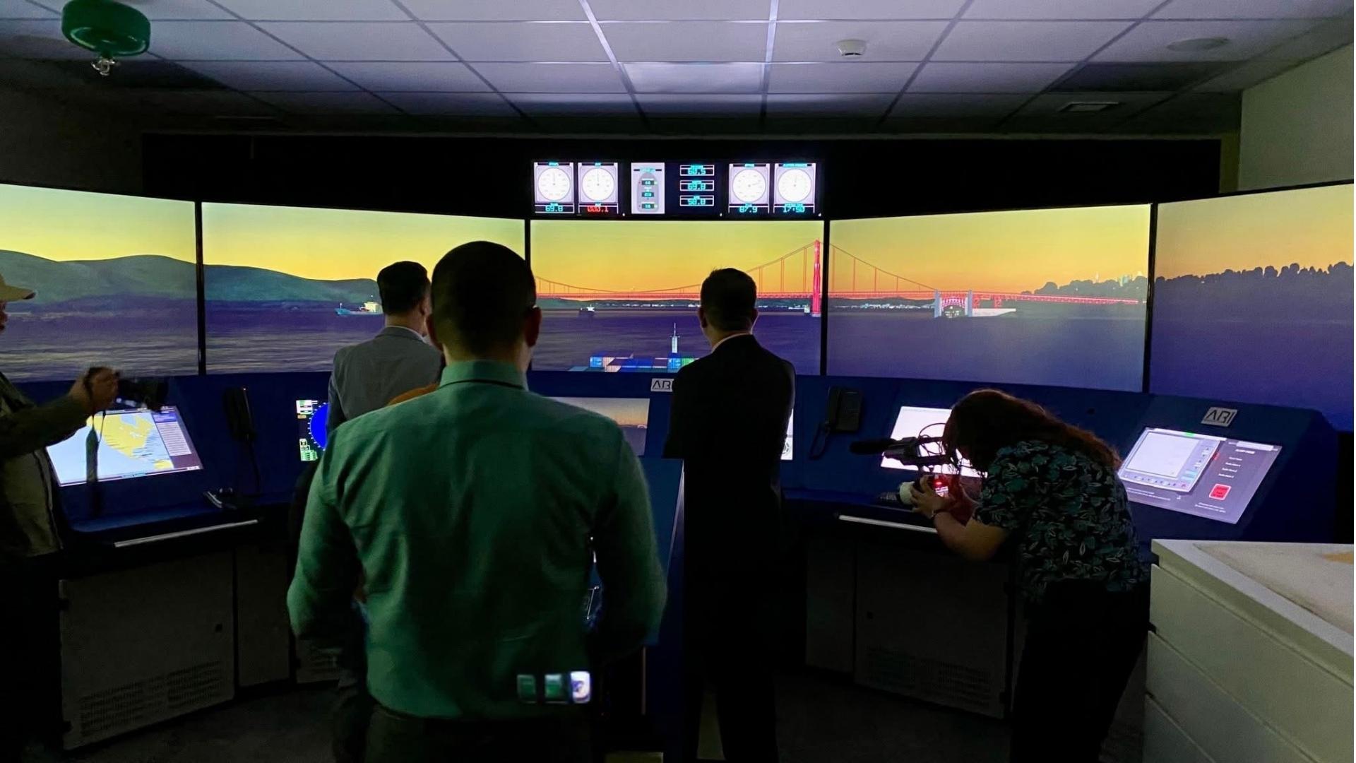 A ship's bridge simulator under a dark blue tint, its windows looking out on a harbour and a suspension bridge
