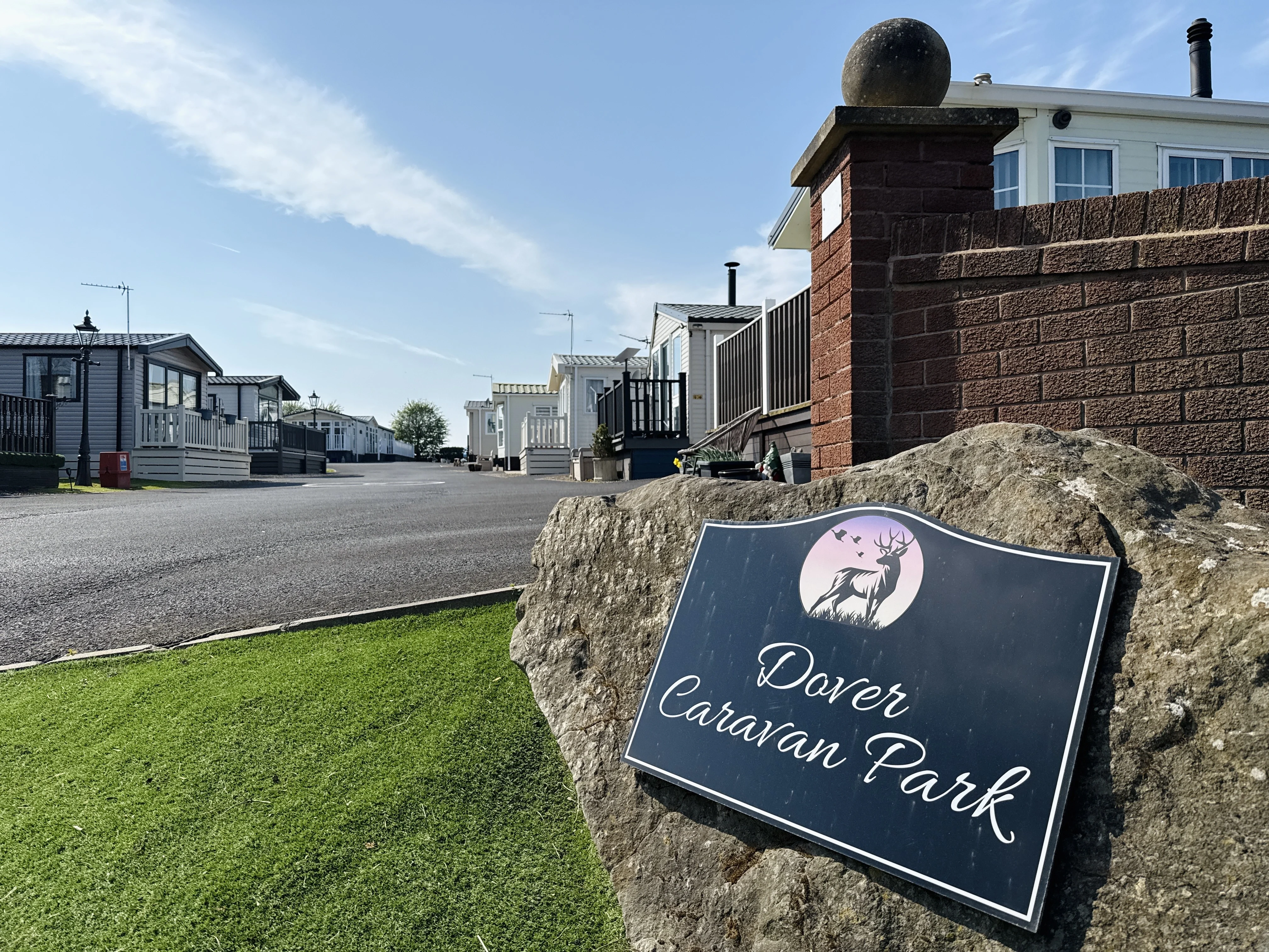 Black sign reading 'Dover Caravan Park' with a deer and birds logo mounted on a large rock near a brick wall and a row of white and grey caravans under a partly cloudy blue sky.