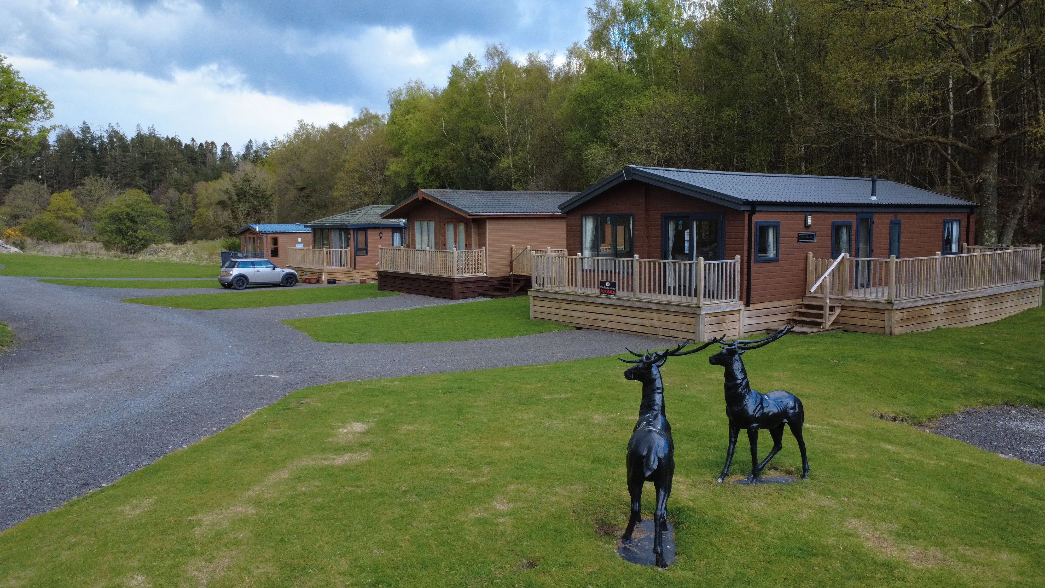 A row of brown wooden cabins with decking in a green grassy area surrounded by trees under a partly cloudy sky, with two black metal deer statues on the grass in the foreground and a silver car parked on a gravel driveway.