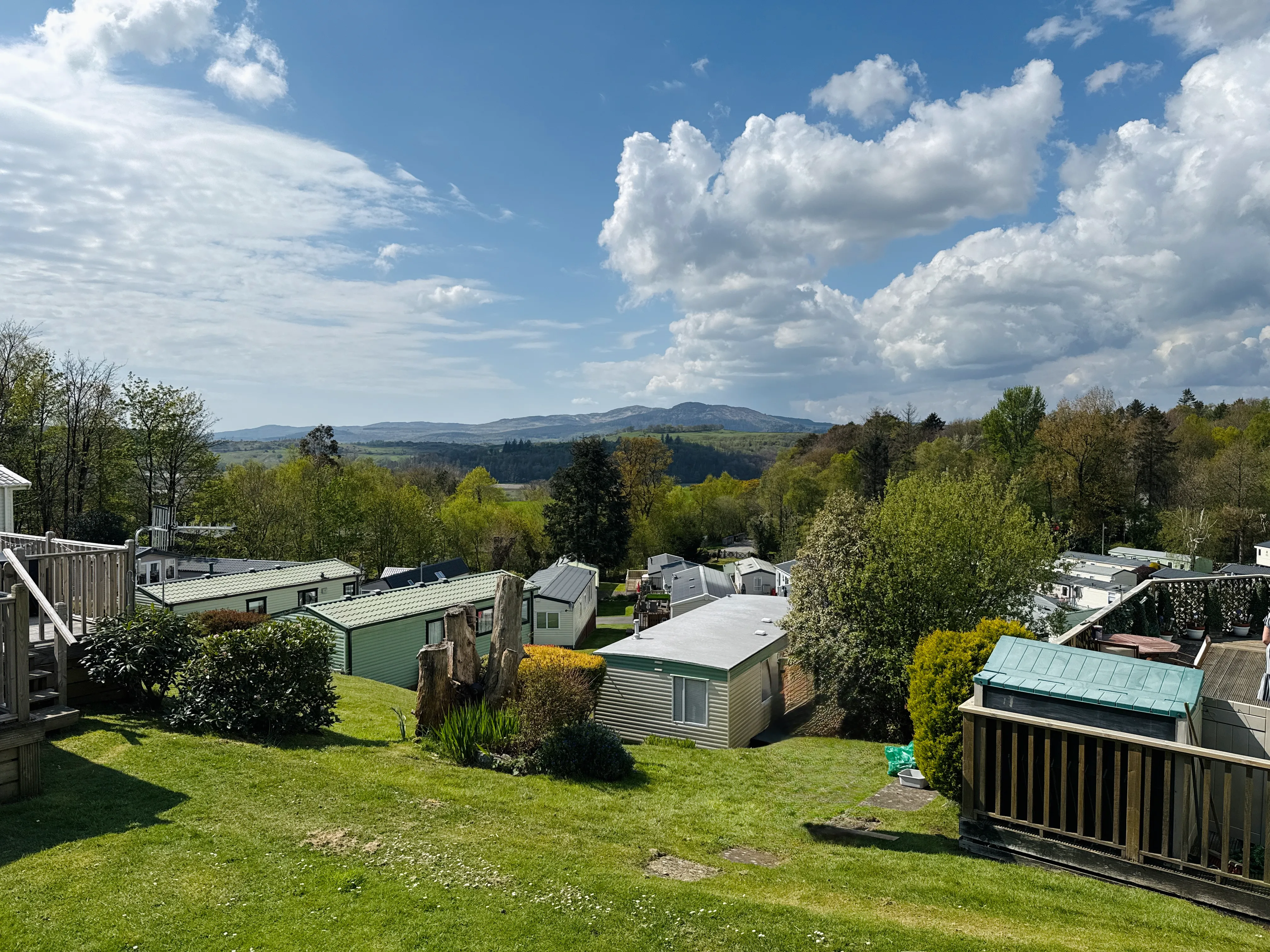 A green grassy area with multiple mobile homes and wooden decks surrounded by trees under a partly cloudy blue sky with hills in the distance.