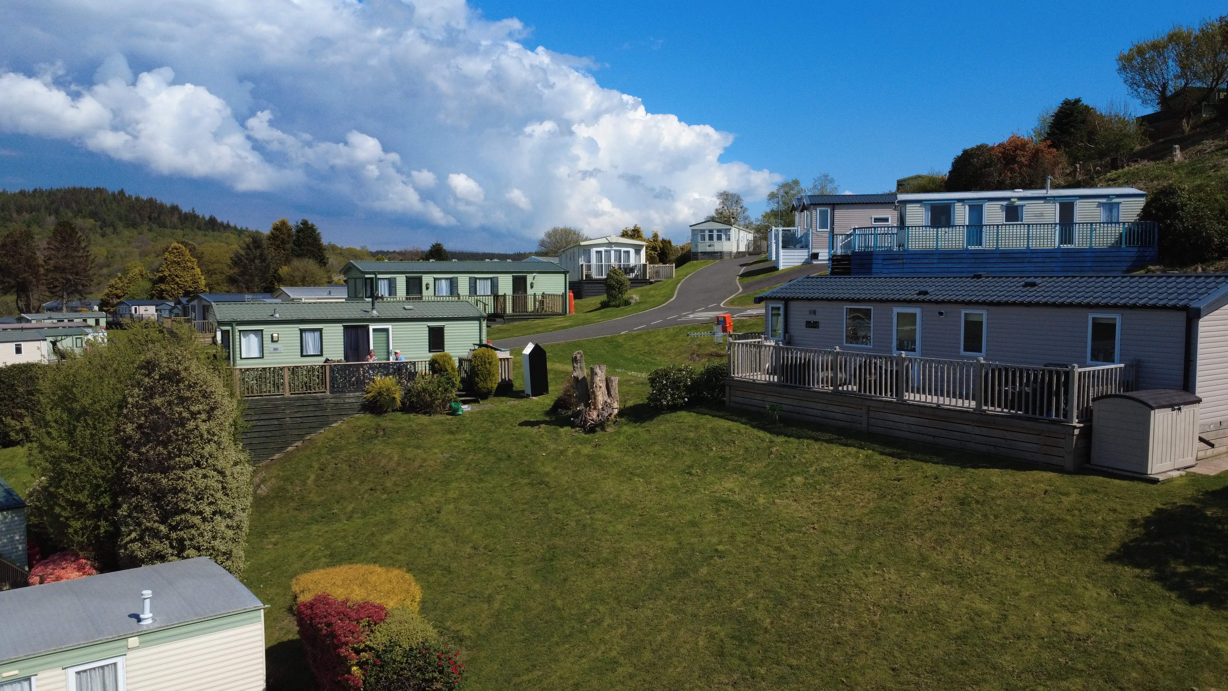 A scenic view of a caravan park with multiple mobile homes arranged on grassy terraces along paved paths under a partly cloudy blue sky, surrounded by trees and hills.