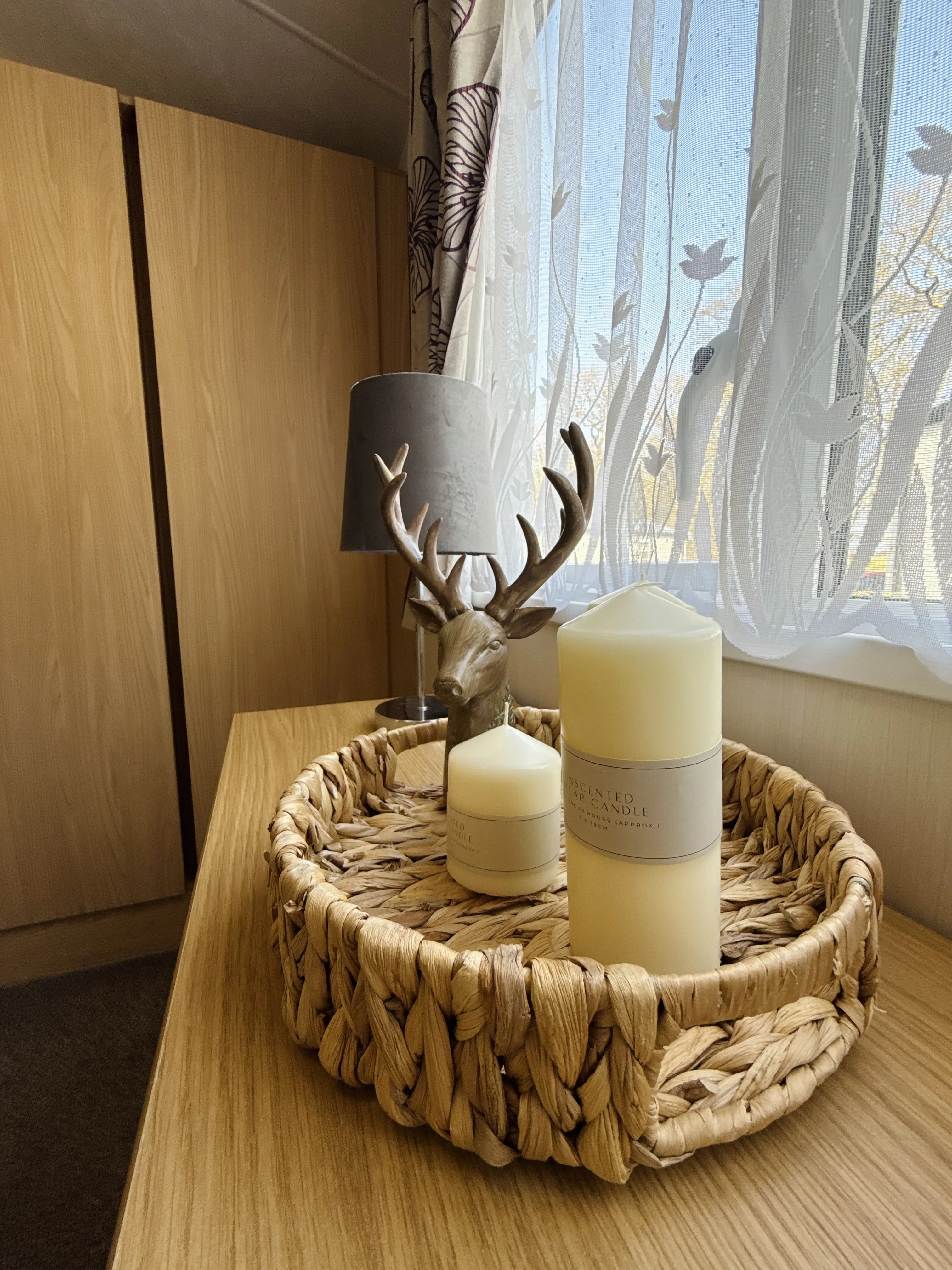A woven basket on a wooden surface holds two ivory scented candles of different sizes and a decorative deer head figurine with antlers, near a window with sheer floral curtains and a grey lampshade in the background.