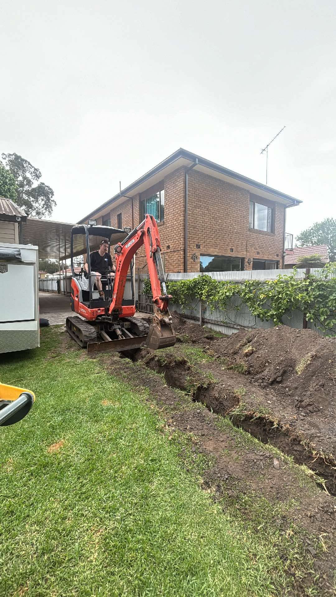 Steele Plumbing mini excavator digging a pipe trench across a backyard lawn beside a brick house