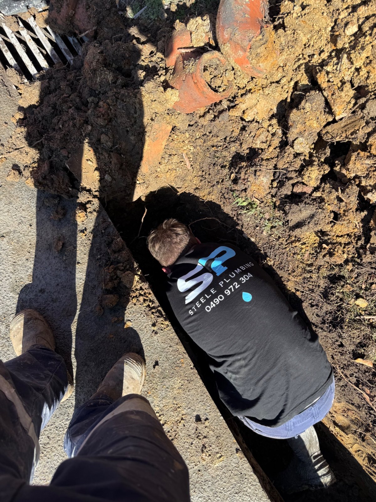 Steele Plumbing plumber in branded shirt working down in a trench beside old terracotta clay pipe
