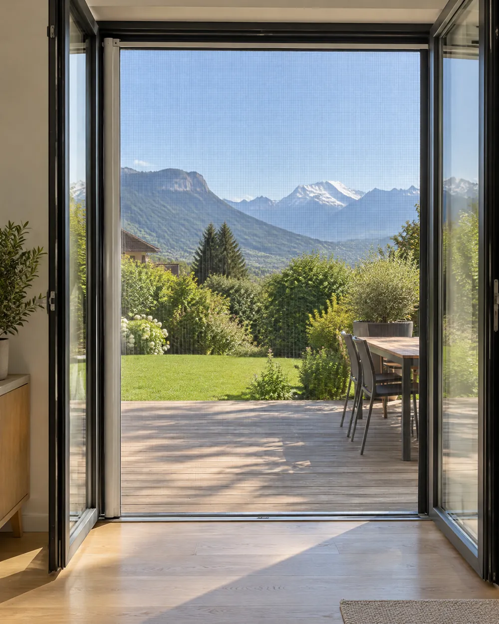 Porte-fenêtre ouverte sur une terrasse en bois, fermée par une moustiquaire latérale à toile presque invisible, montagnes au loin.